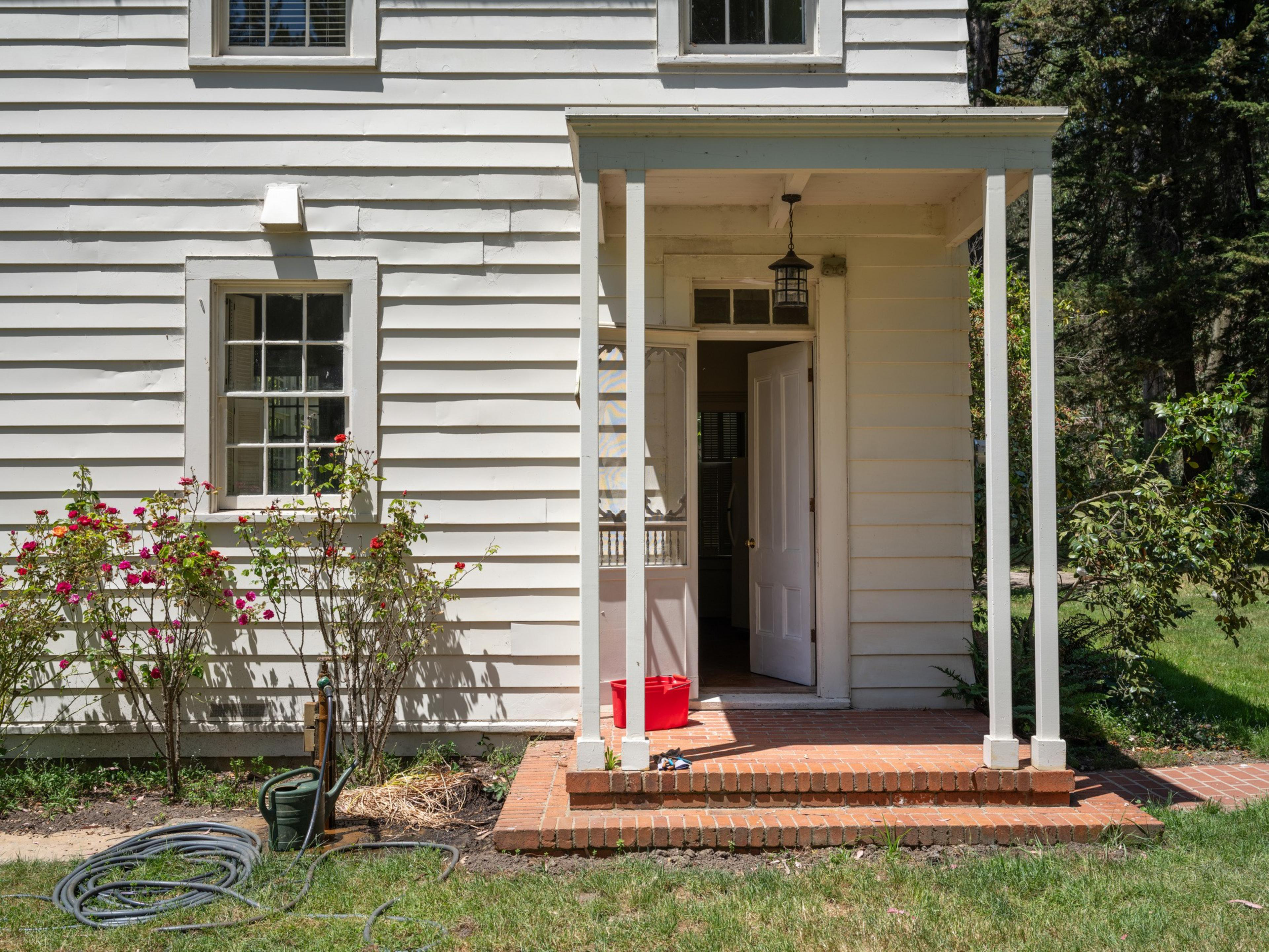 A white wooden house features an open door on a small porch with two columns. A garden hose and watering can are on the grass, near blooming red roses.