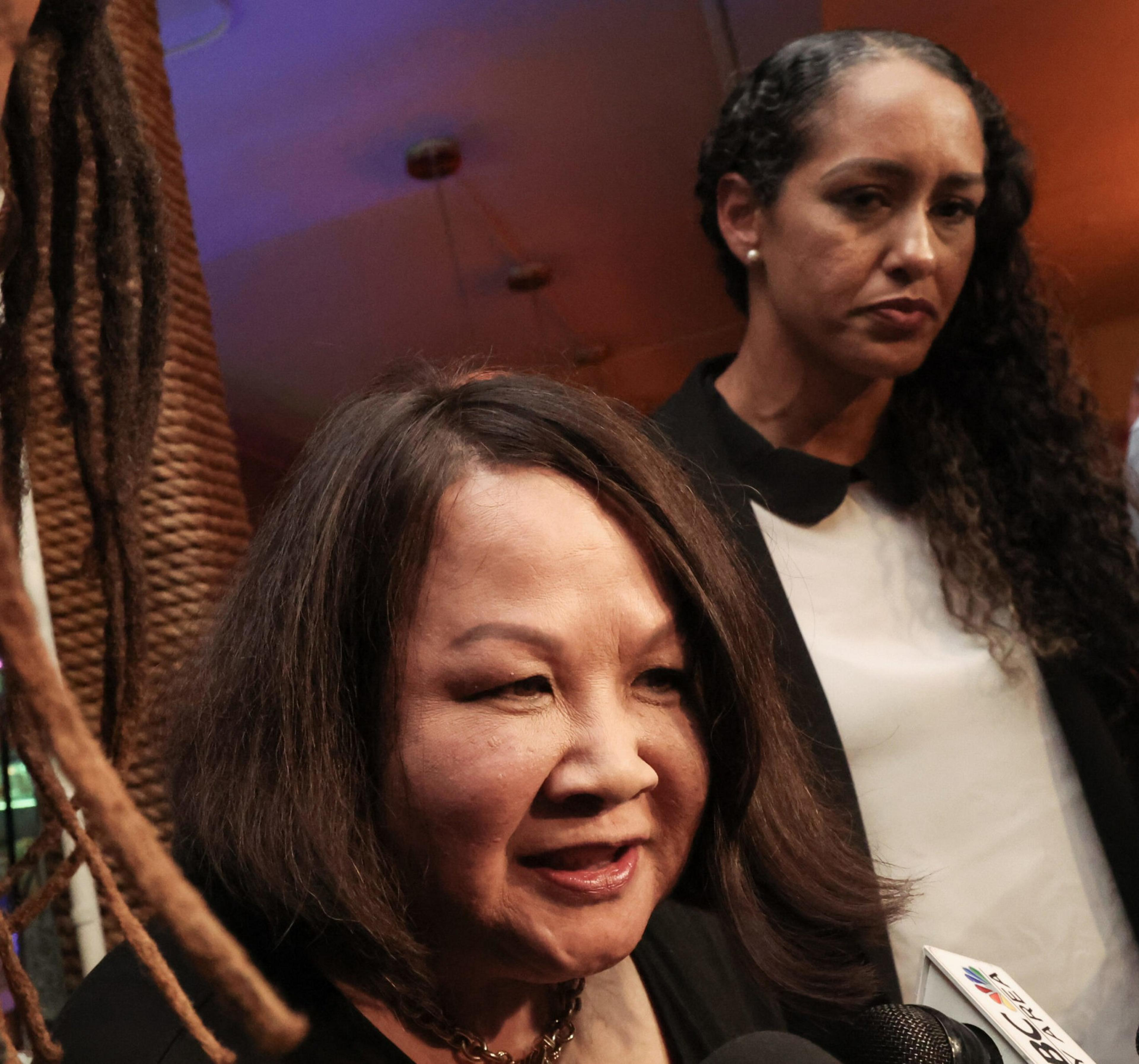 A woman with shoulder-length dark hair speaks into microphones while another woman with long curly hair and a serious expression stands behind her.
