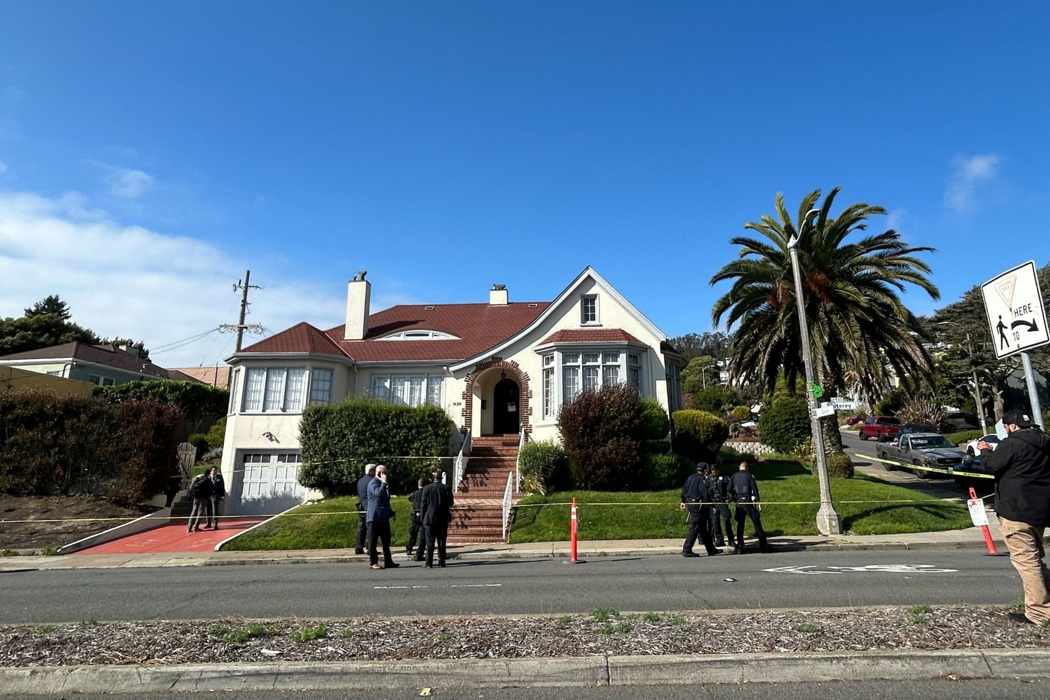 A two-story house with a red roof and white exterior sits behind trimmed bushes and a brick staircase, while several people and police officers stand outside.
