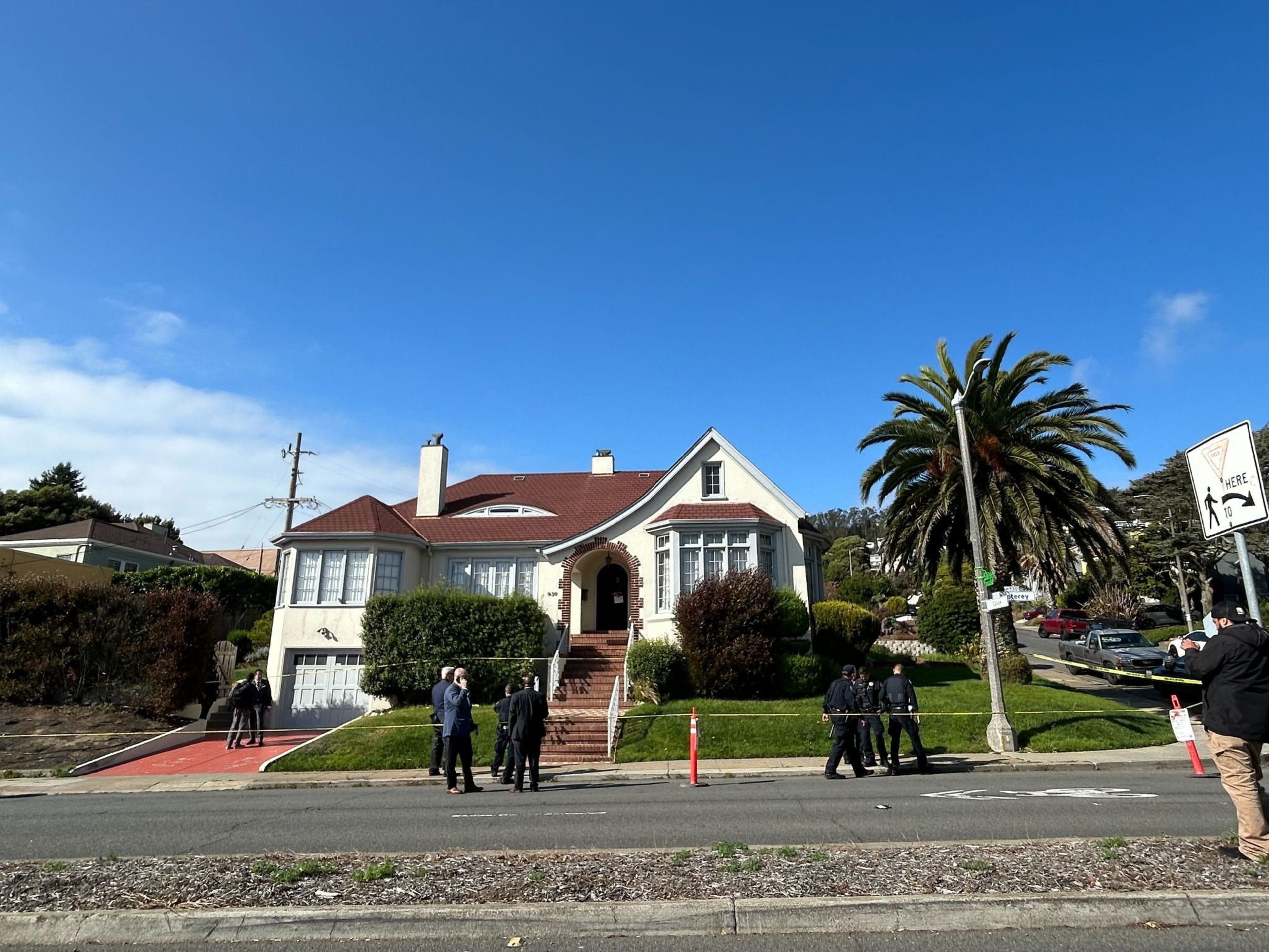 A two-story house with a red roof and white exterior sits behind trimmed bushes and a brick staircase, while several people and police officers stand outside.
