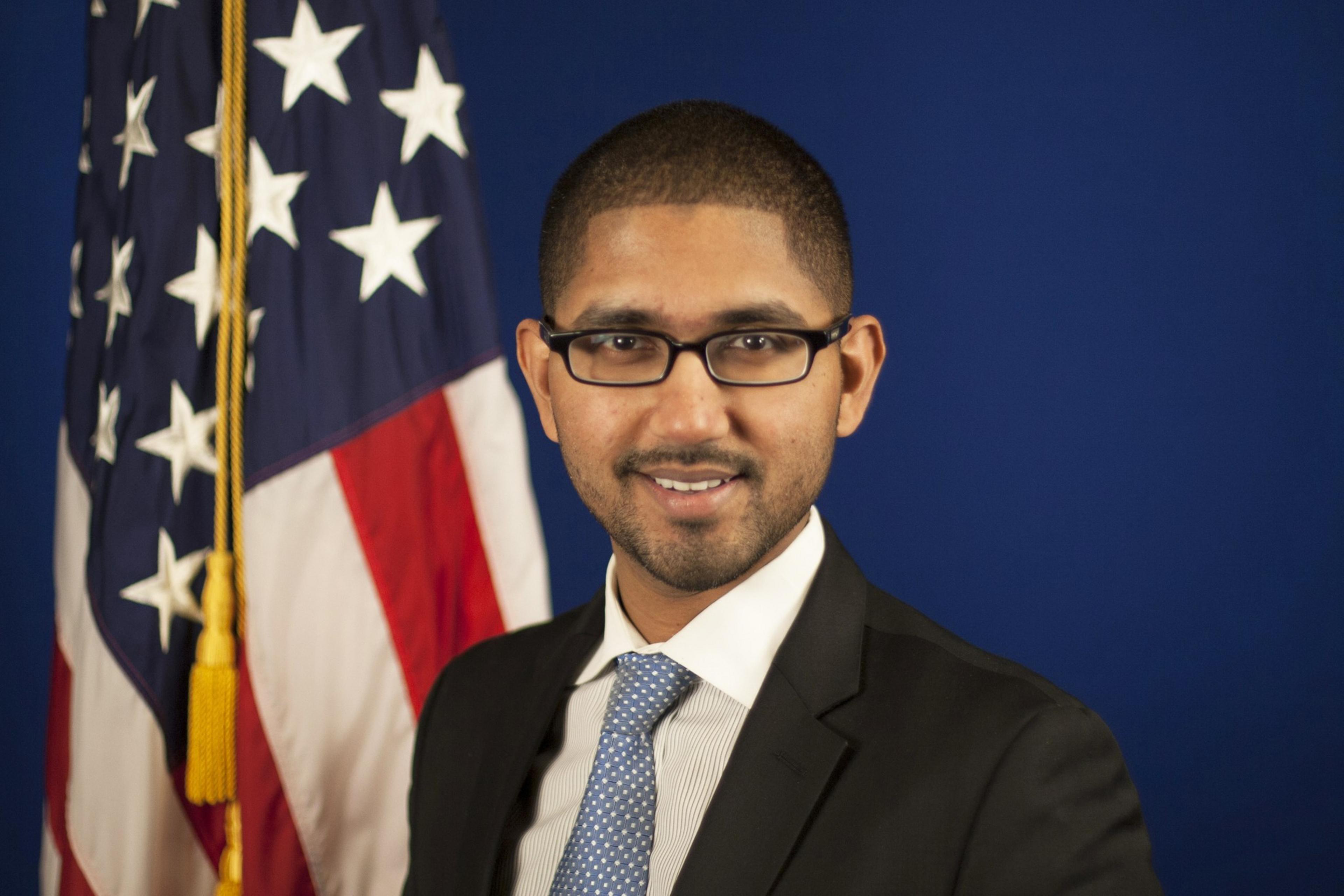 A man wearing glasses, a dark suit, white shirt, and blue polka dot tie smiles in front of an American flag and a blue background.