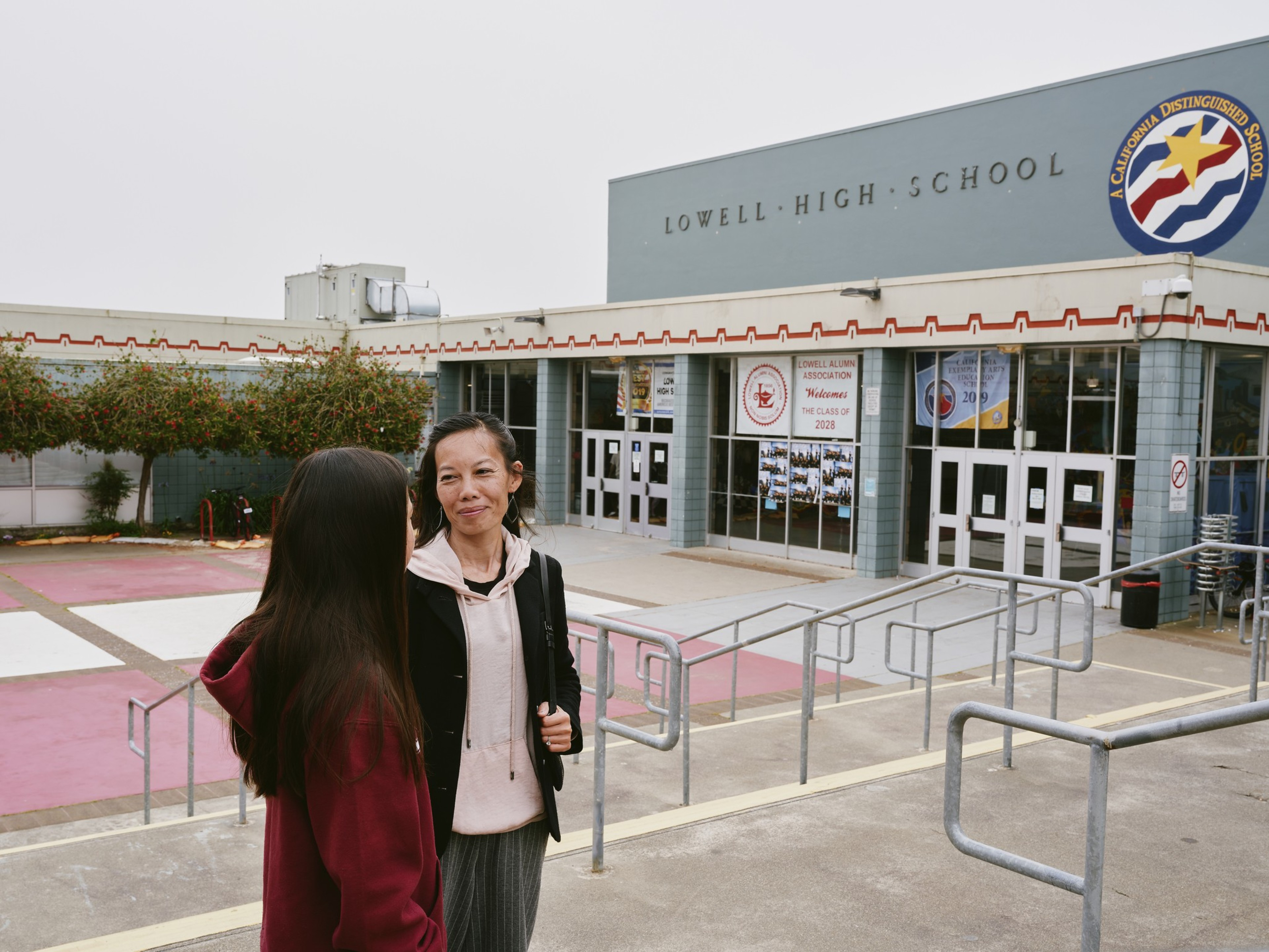 Two people stand outside Lowell High School. The building has a sign for a California Distinguished School and posters for the Class of 2028.