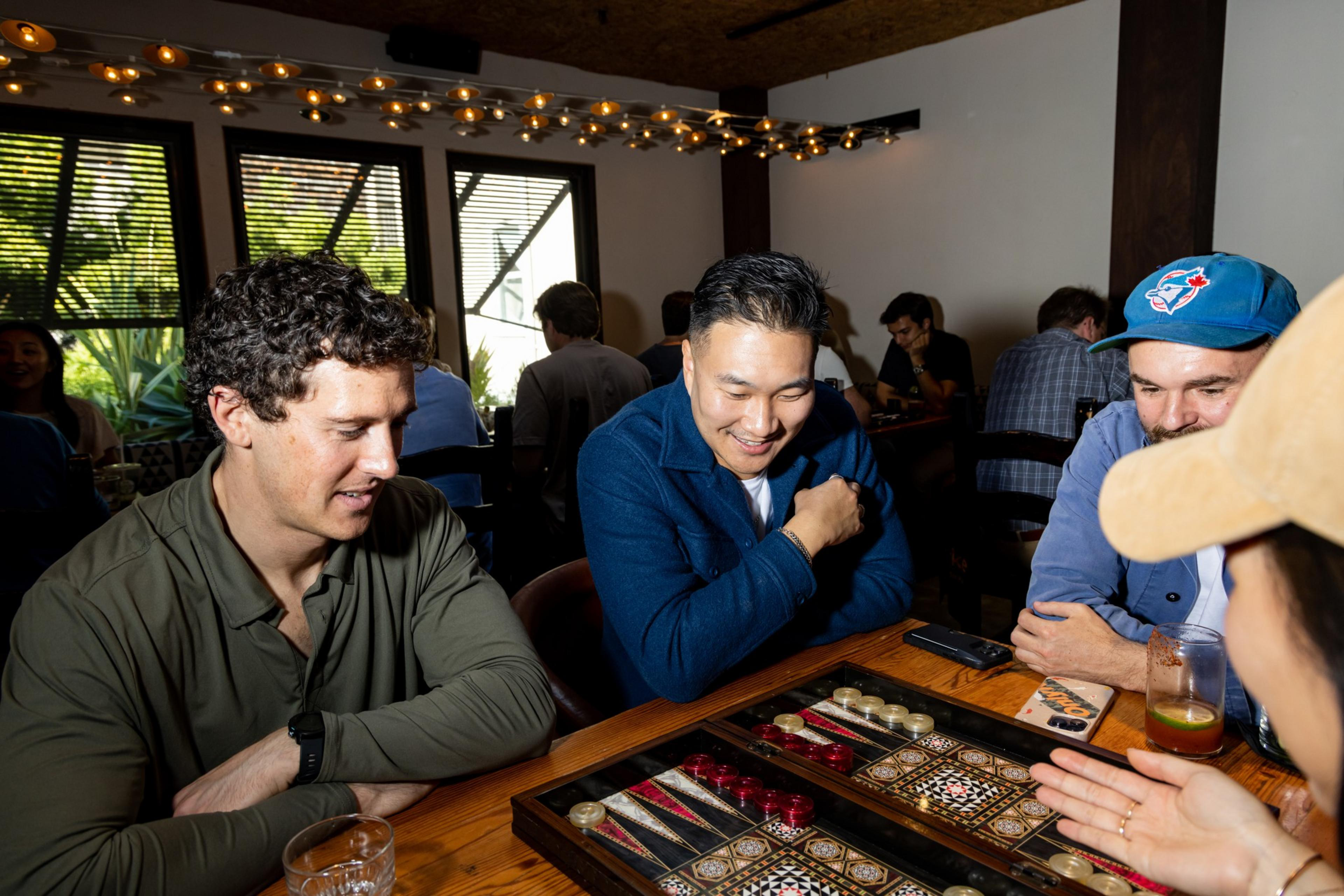 Three men sit around a wooden table smiling and concentrating on a backgammon game while a fourth person gestures towards the board.