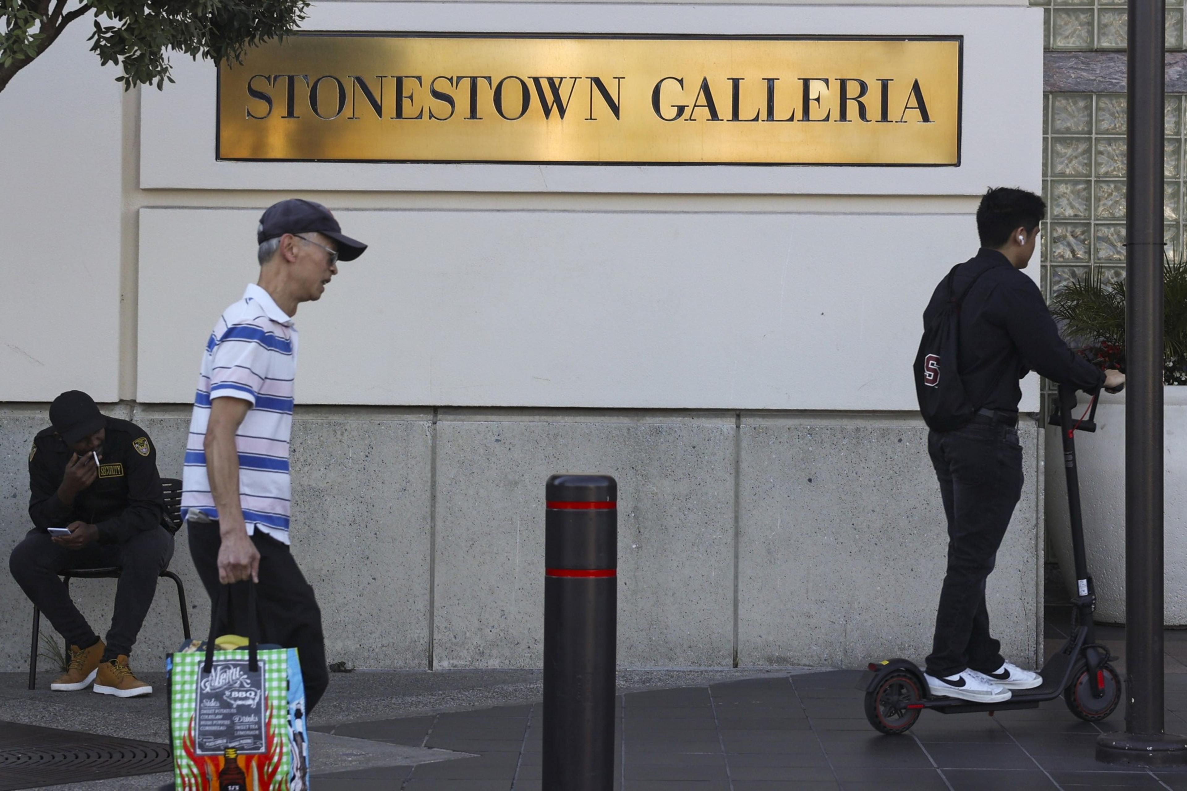 Two shoppers walk past a brass plaque that reads, "Stonestown Galleria."