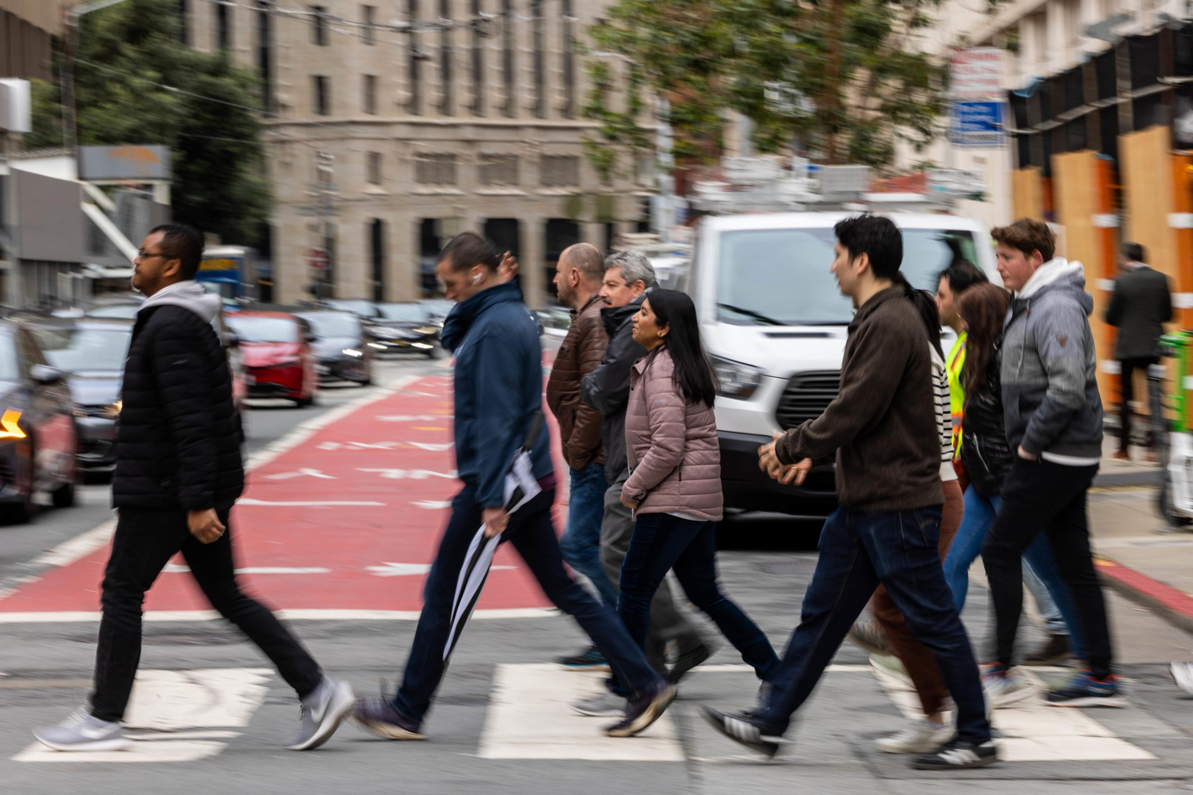 A group of people crosses a city street at a crosswalk. They walk briskly, while cars are stopped. A red bus lane and buildings are visible in the background.