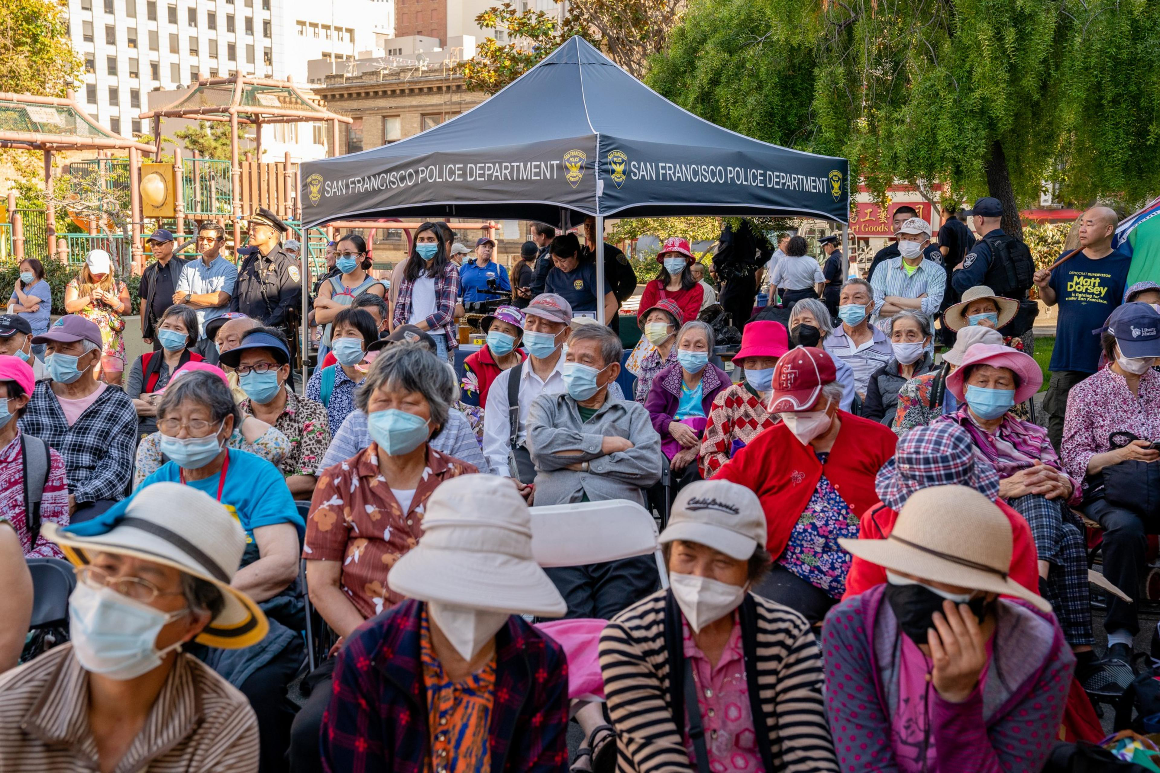 A crowd of people, mostly elderly with masks, sitting outdoors with a police tent in the background.