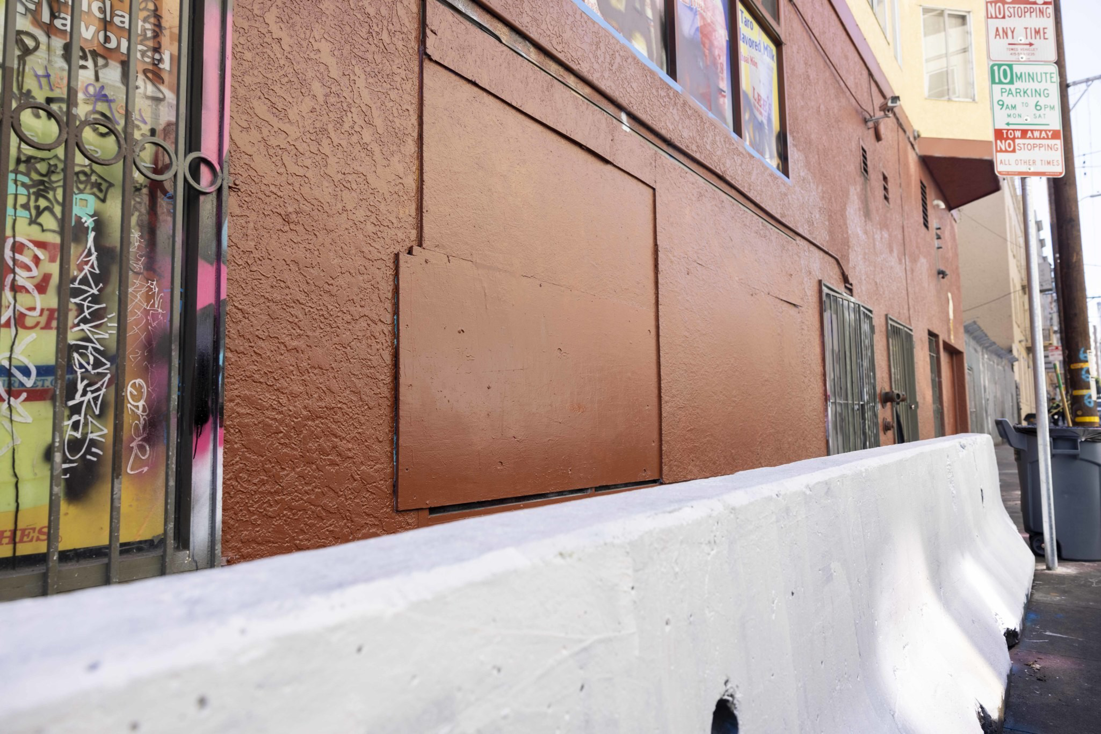 A textured brown wall with boarded sections, graffiti-tagged window, metal bars, a white concrete barrier, and parking signs in an urban alley.