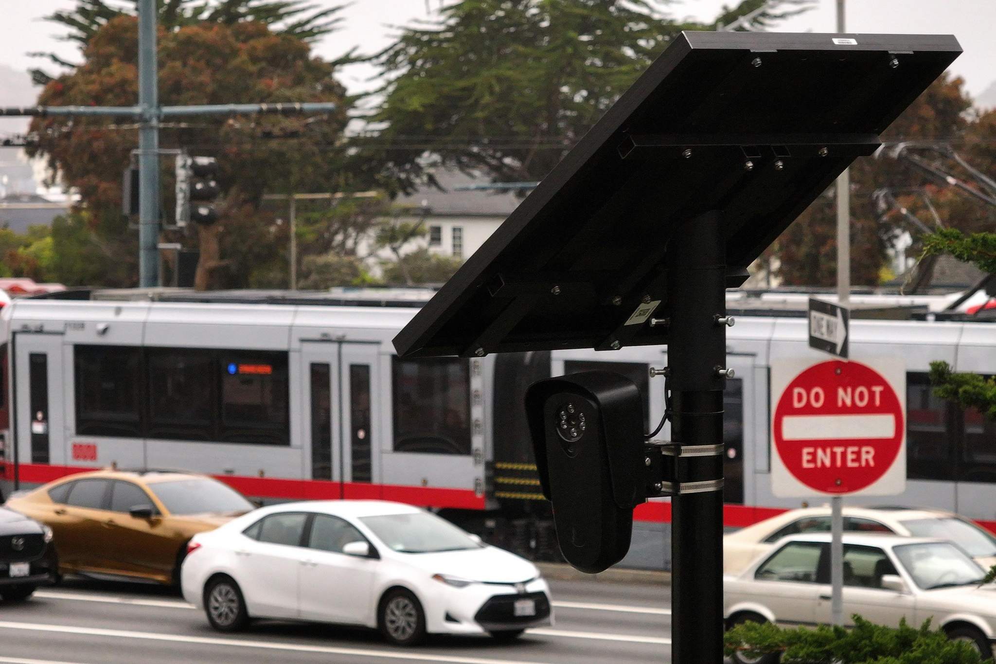 The image shows a street scene with a "Do Not Enter" sign, several cars, and a moving tram. A pole with a solar panel and security camera is prominent.
