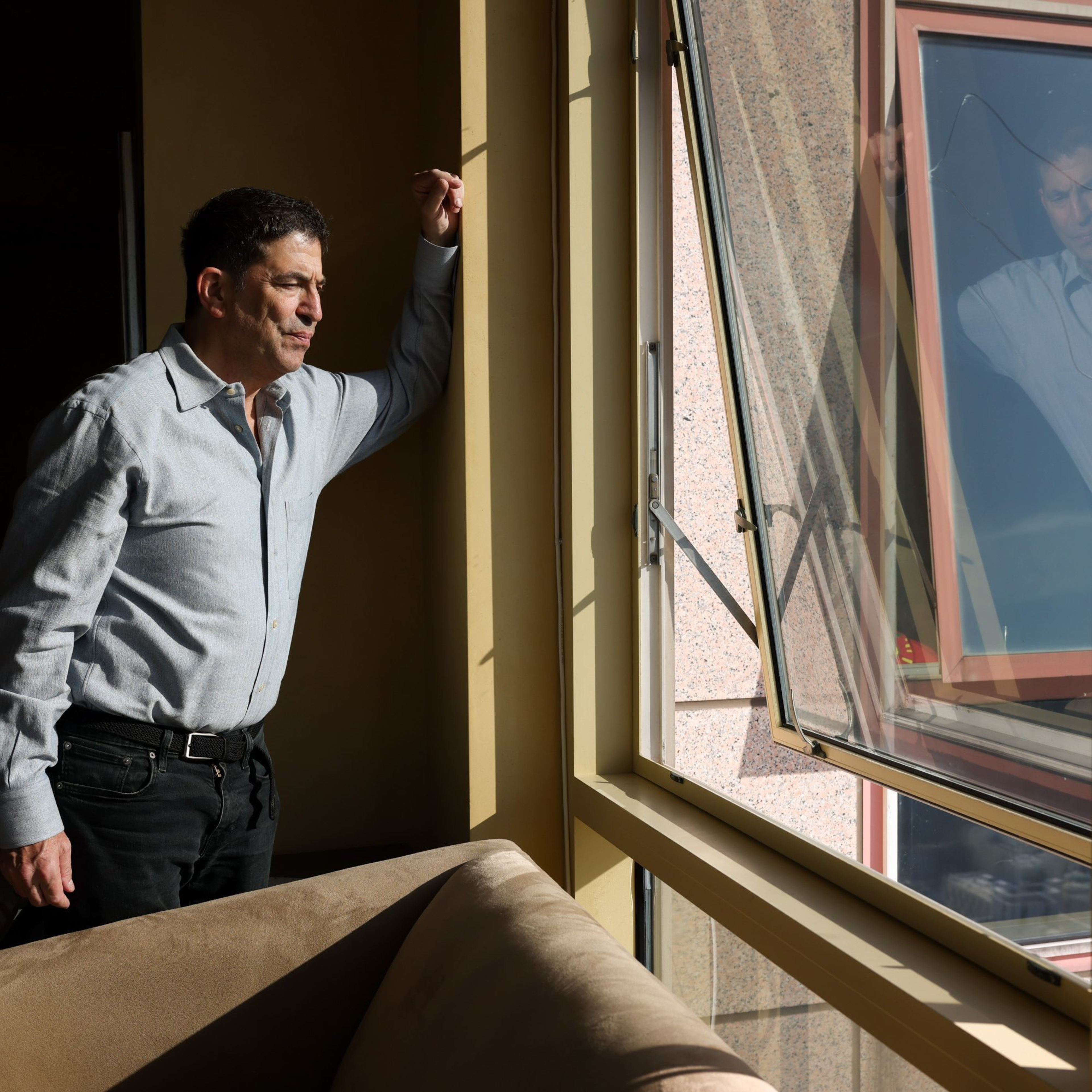 A man in a light shirt stands by an open window, looking outside. Sunlight casts shadows inside the room, and his reflection is visible in the window glass.