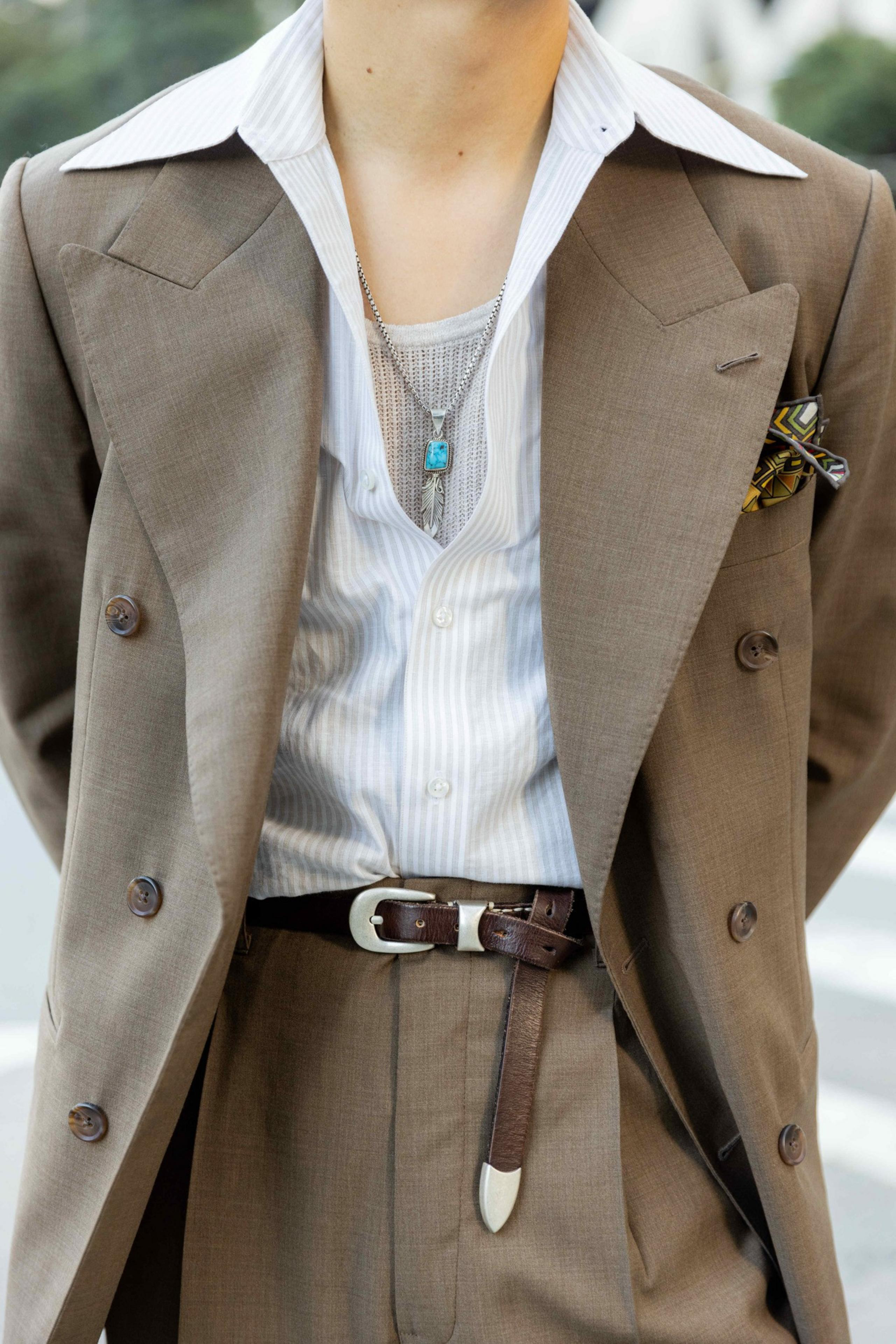A person wears a brown double-breasted suit with a striped shirt, a silver chain necklace with a turquoise pendant, a brown belt, and a patterned pocket square.