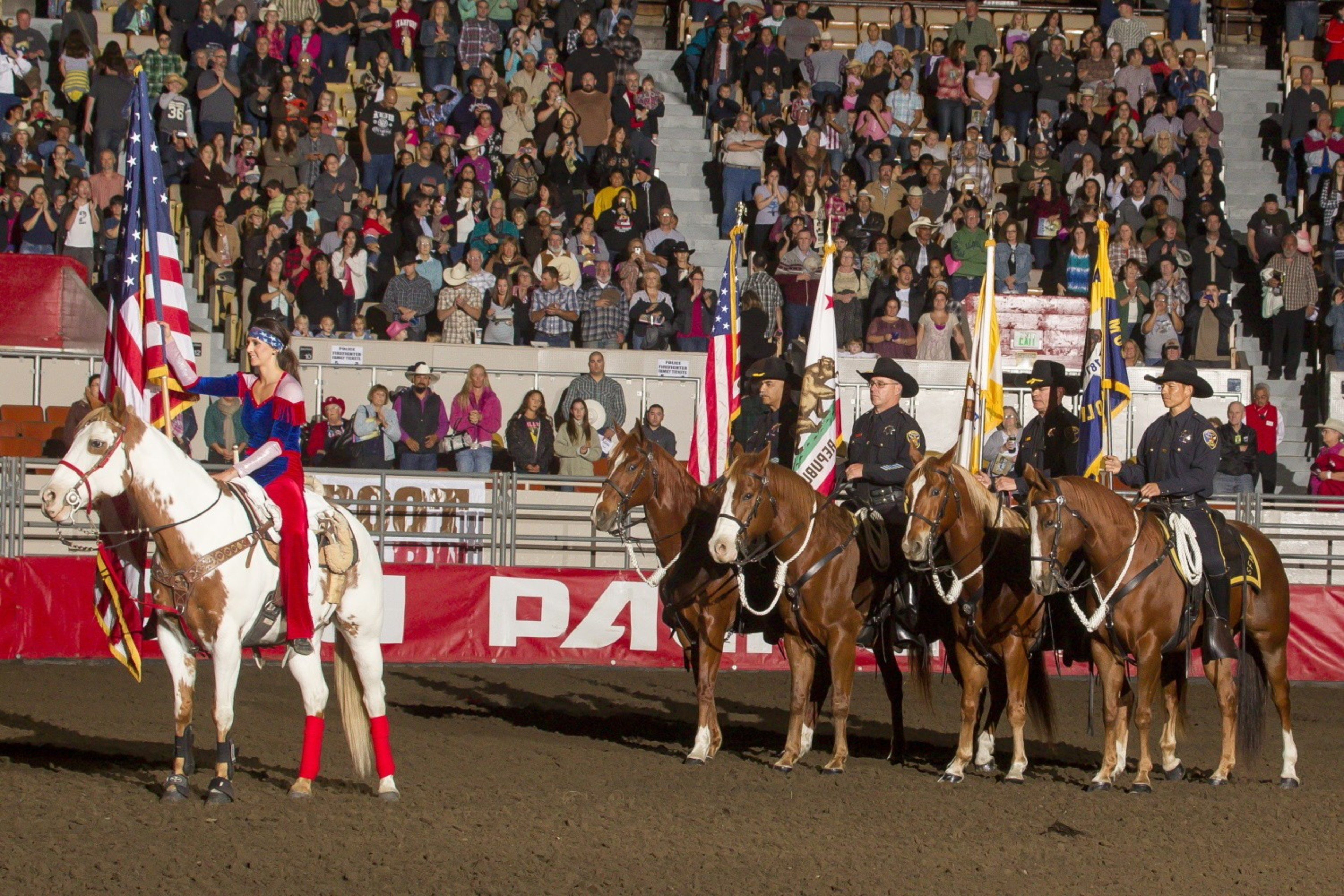 Riders on horseback carry flags in an arena, with spectators watching in the background.