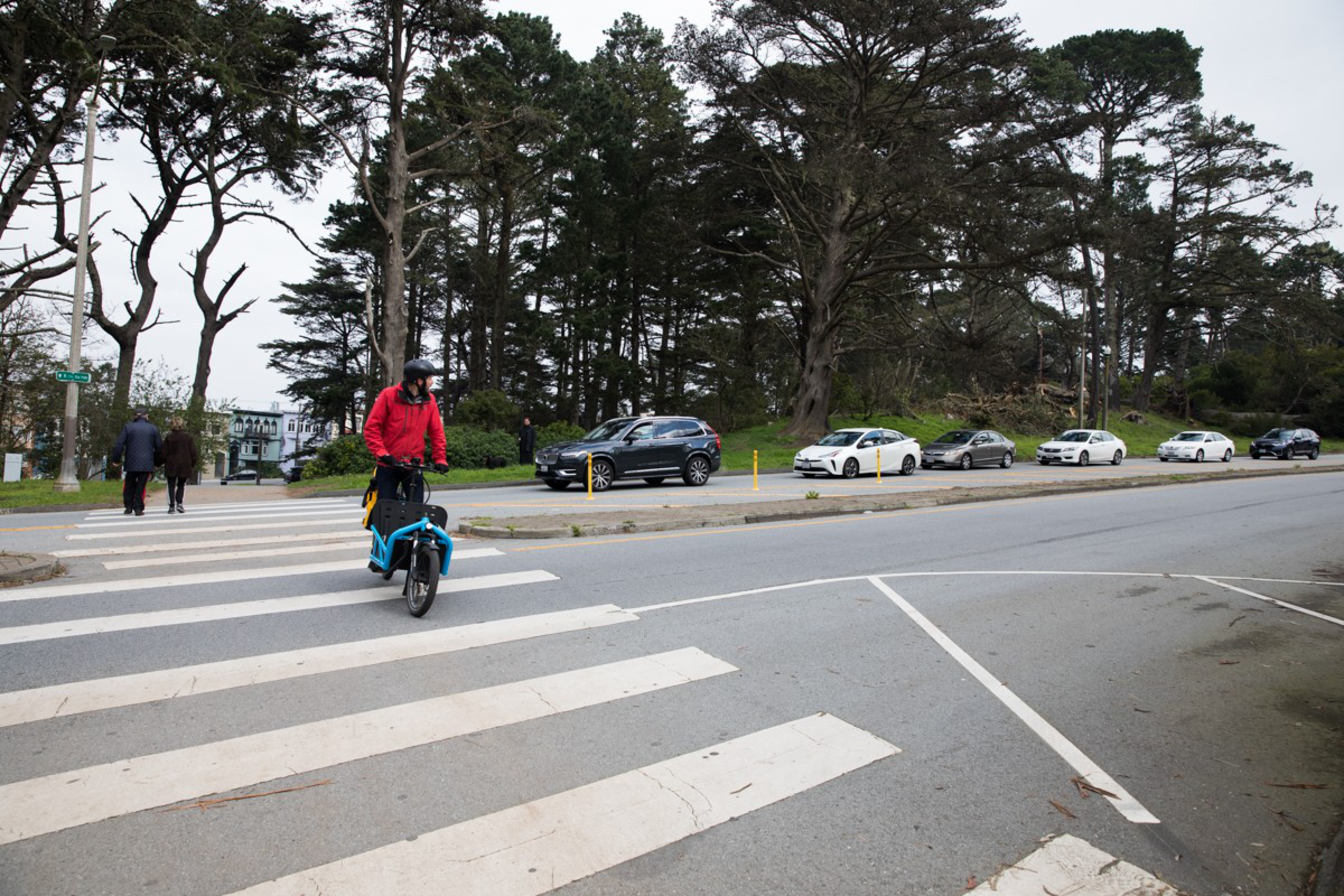 A cyclist in a red jacket rides across a crosswalk on a quiet street lined with parked cars and tall trees. Two people walk in the background.