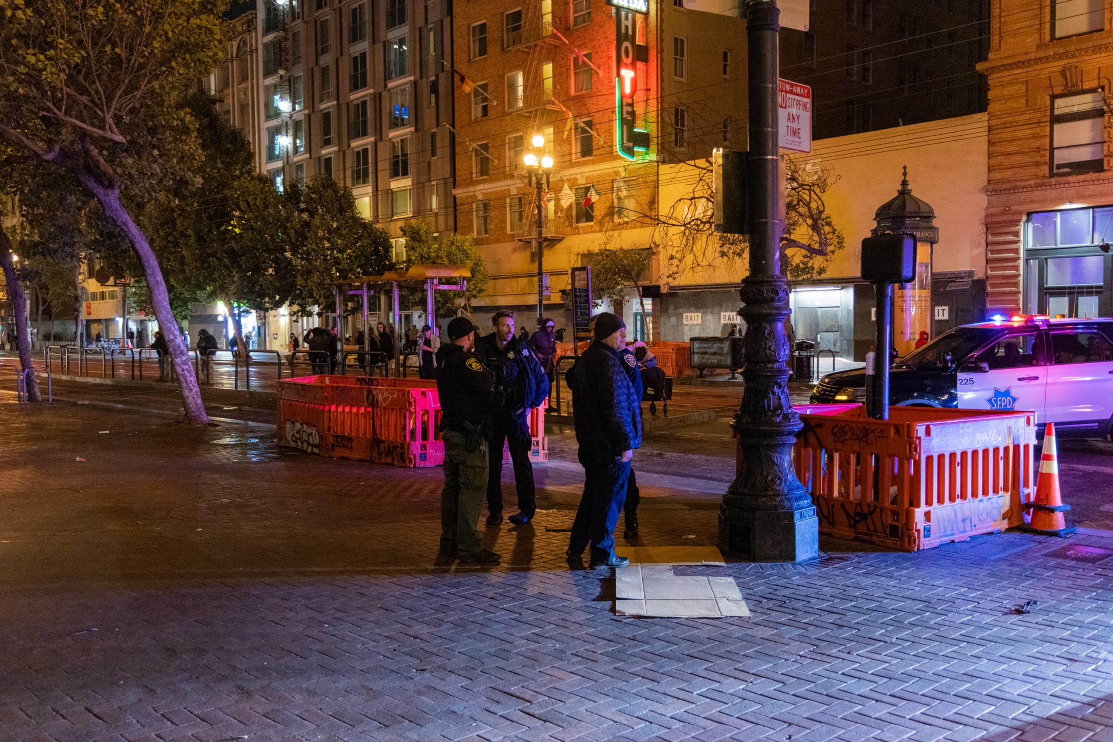 The image shows a night scene with several police officers standing next to orange barricades and a police SUV with flashing lights, in a city setting with tall buildings.