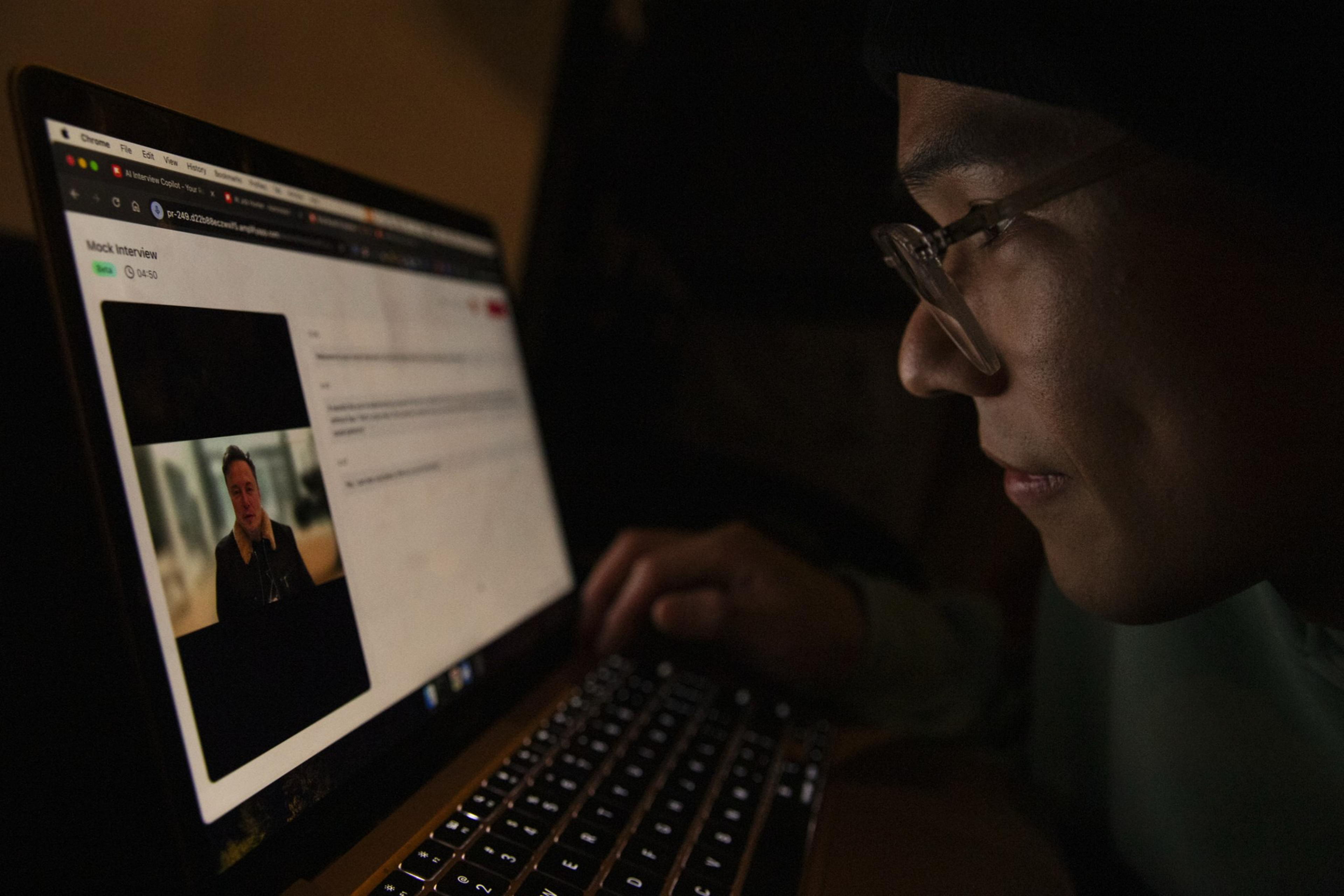 A person wearing glasses is intently working on a laptop displaying a mock interview video with a man speaking.