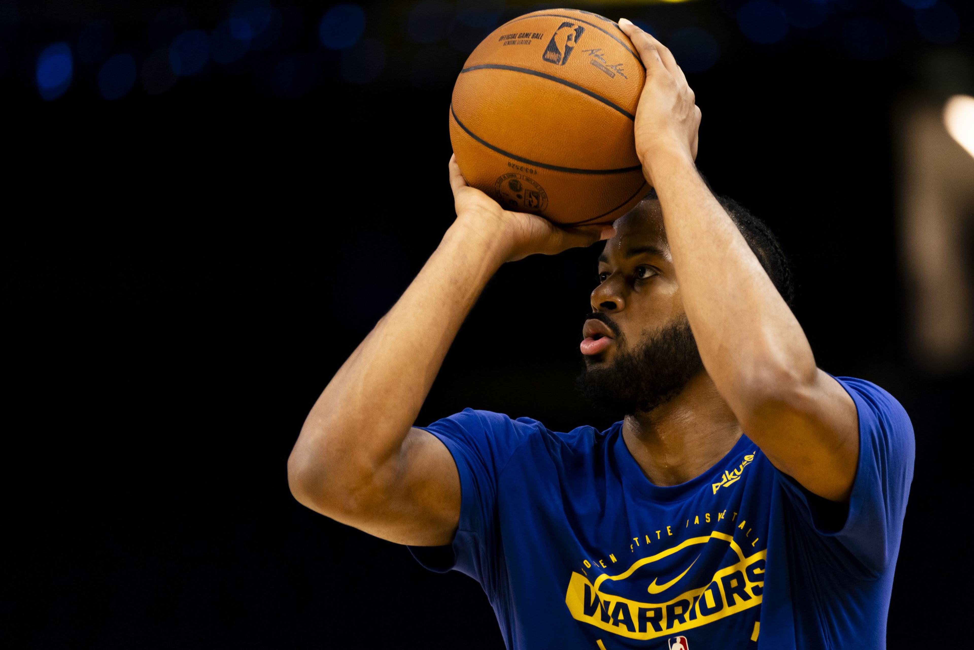 A basketball player wearing a blue Golden State Warriors shirt prepares to shoot, holding an NBA basketball above his head with both hands.
