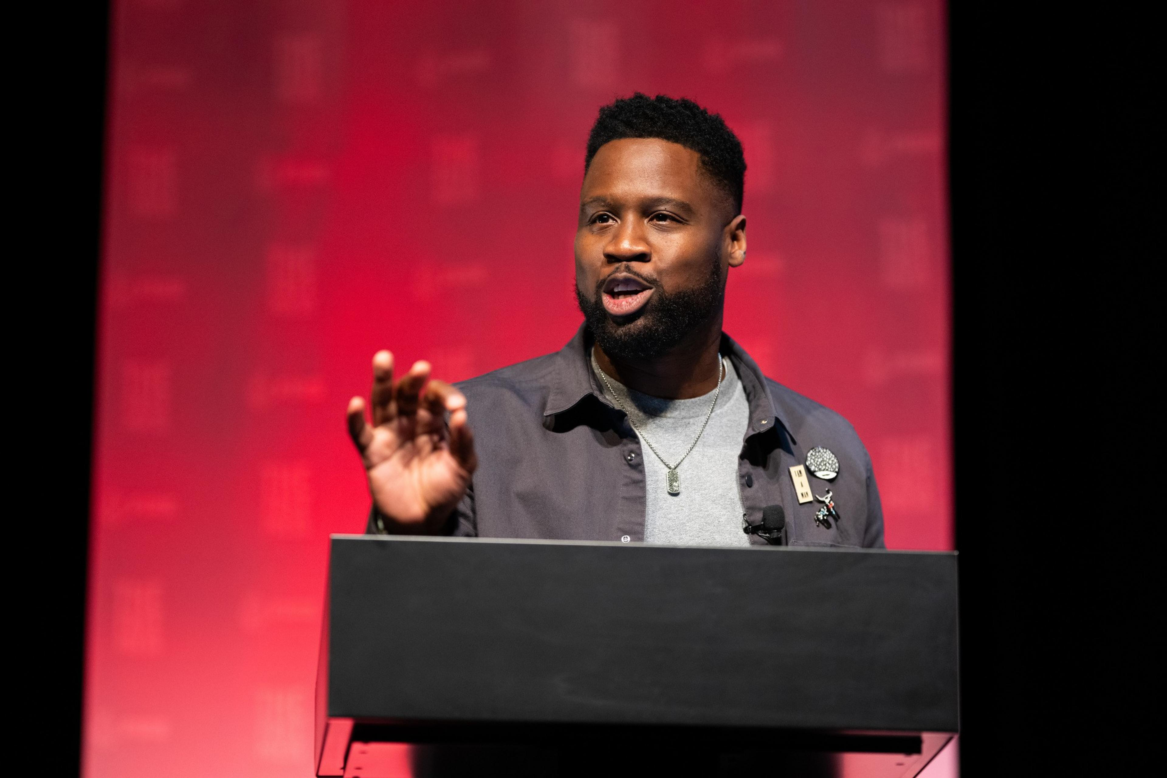 A man stands speaking at a podium with a red background, wearing a grey jacket adorned with pins over a light shirt, gesturing with his right hand and looking engaged.