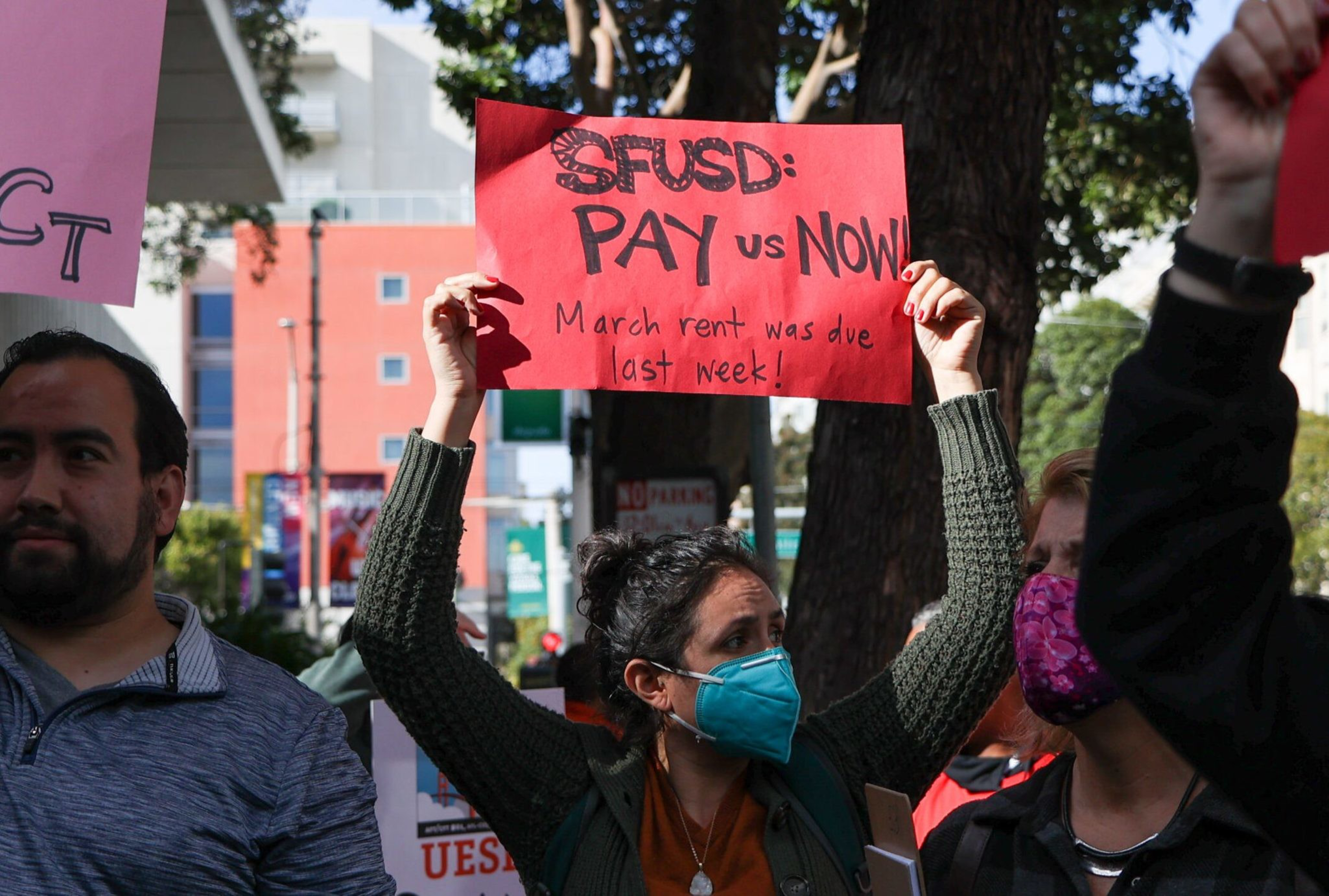 A group of people are holding signs in a protest. One woman is raising a red sign that reads, "SFUSD: PAY us NOW! March rent was due last week!" They're outdoors with trees in the background.