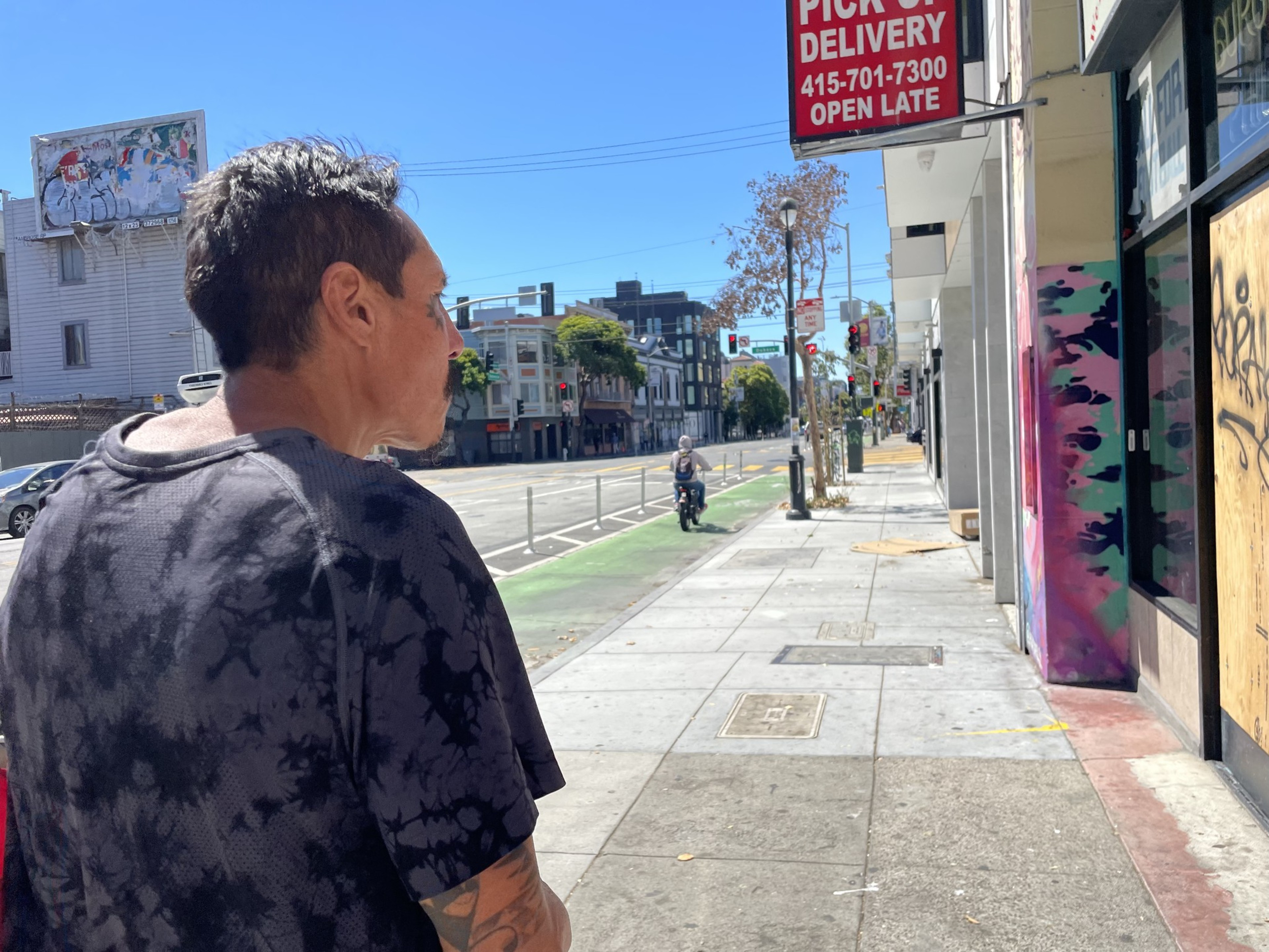 A man with short hair and a dark, tie-dye shirt is walking on a sunny city street with a bike lane and a &quot;Pick Up &amp; Delivery&quot; sign in the background.