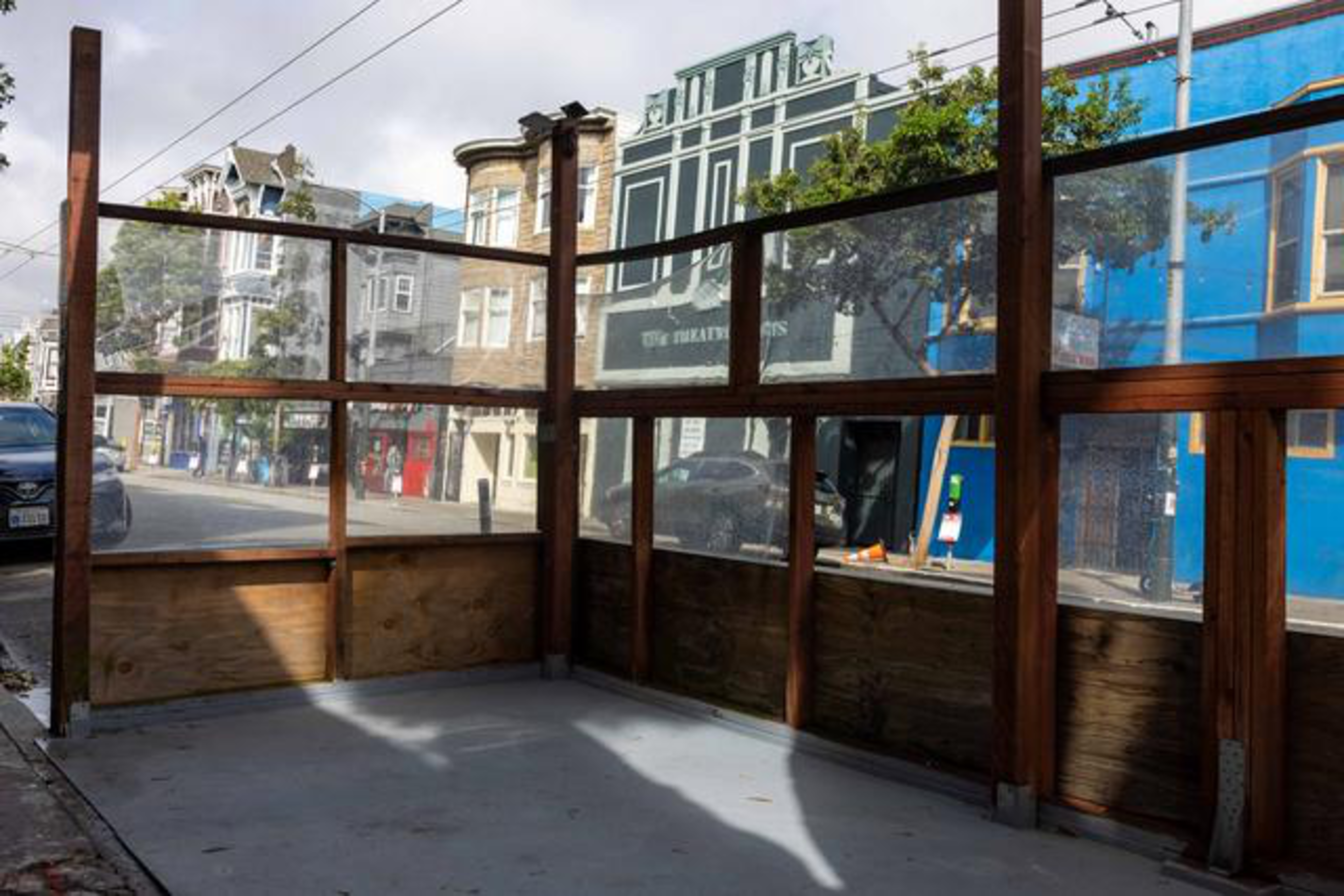 A wooden and glass outdoor enclosure sits on a sidewalk with colorful buildings and cars visible on the street behind it.