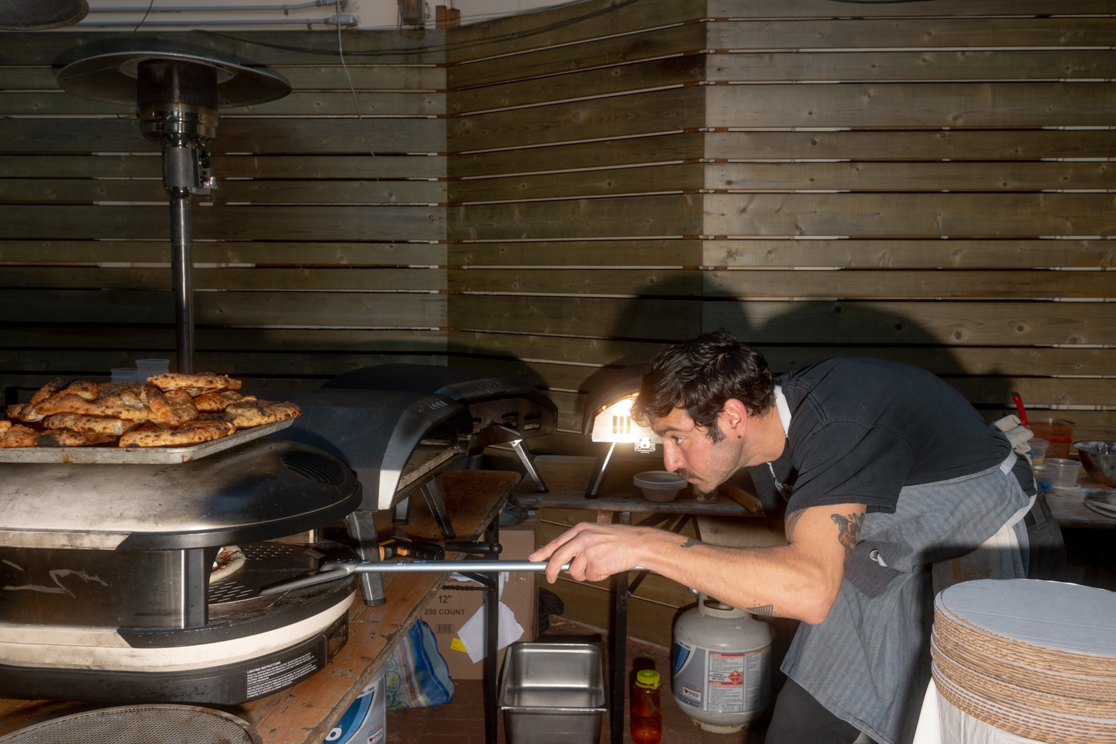 A person in a black shirt and apron is focused on baking pizza in an outdoor oven. A tray of cooked pizzas is nearby, and a wooden wall forms the background.