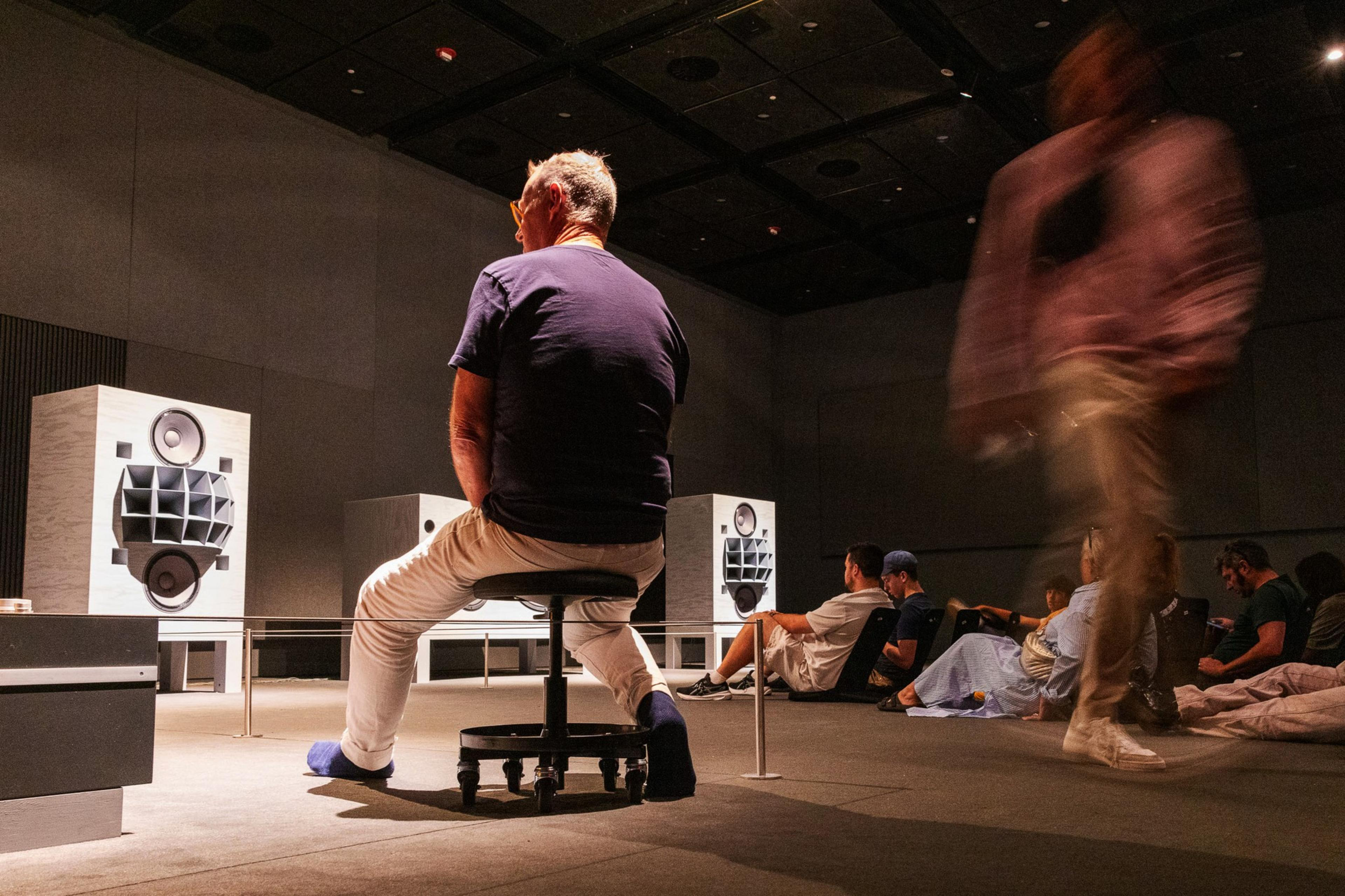 A man sits on a stool in a dimly lit room with large speakers. Other people are seated or walking around, with some blurred motion indicating movement.