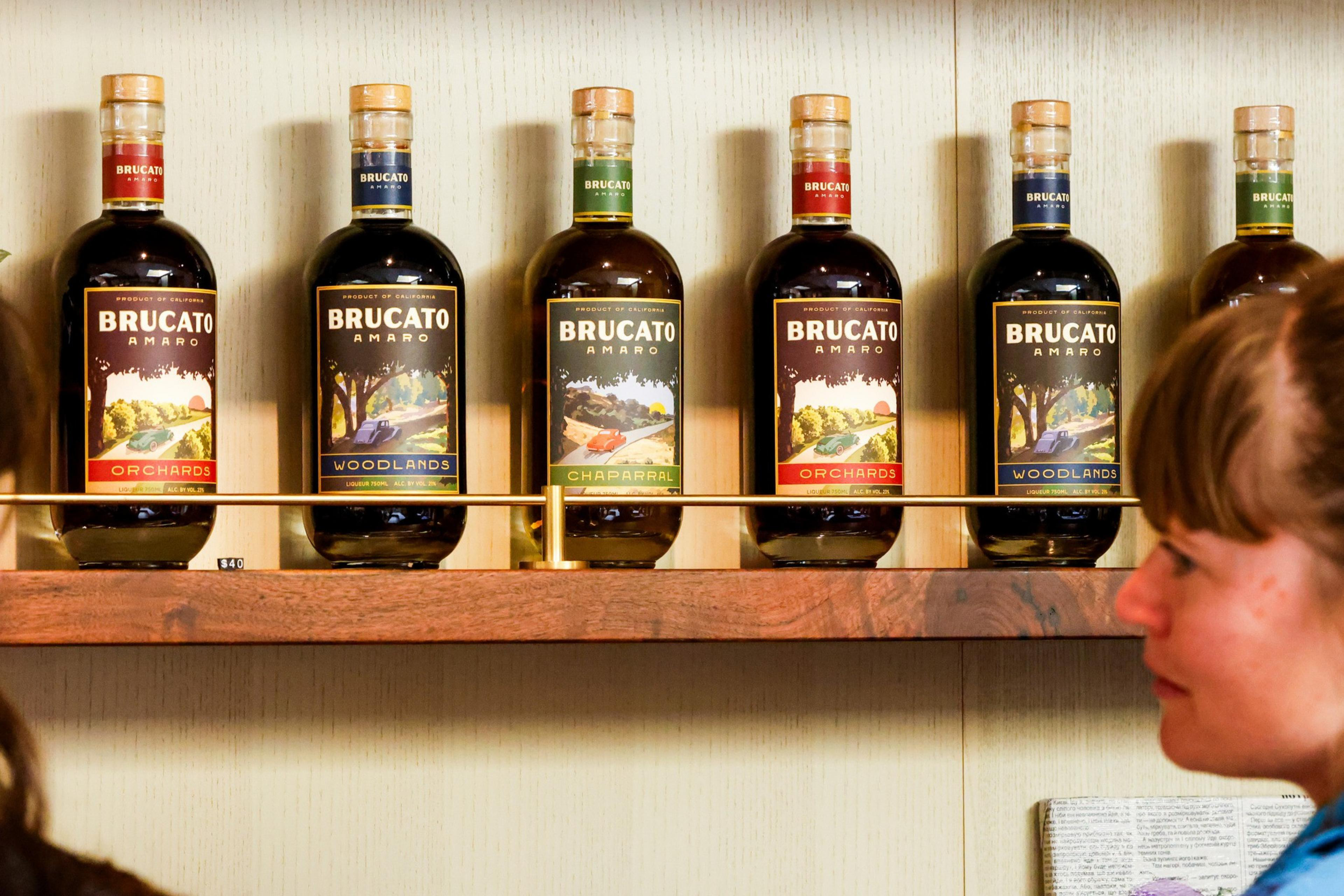 This image shows six bottles of Brucato Amaro on a wooden shelf, each featuring a different label design. There are two people partially visible in the foreground.