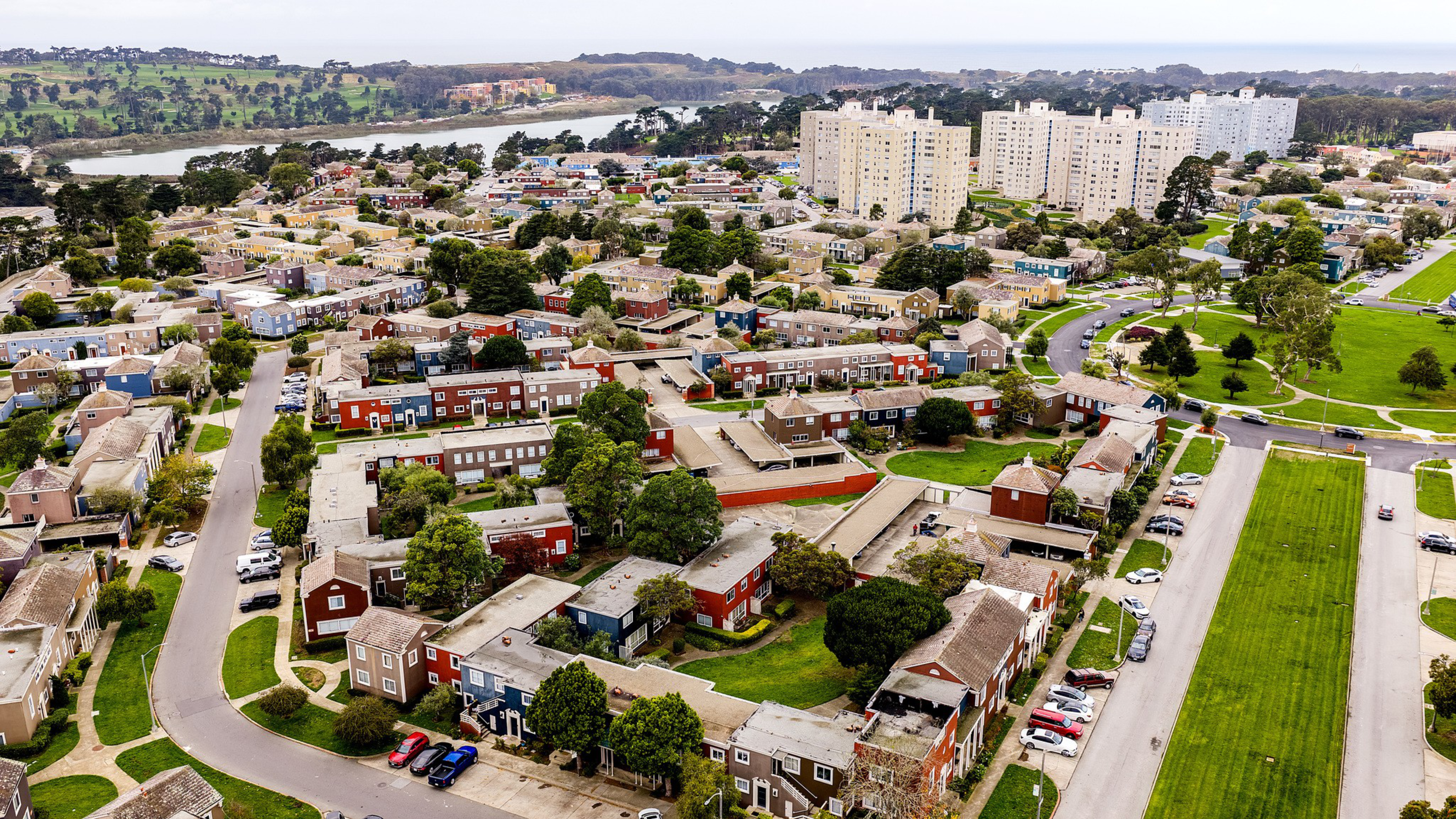 A residential area features colorful townhouses with green lawns, surrounded by trees, parked cars, winding roads, high-rise buildings, and a water body in the distance.