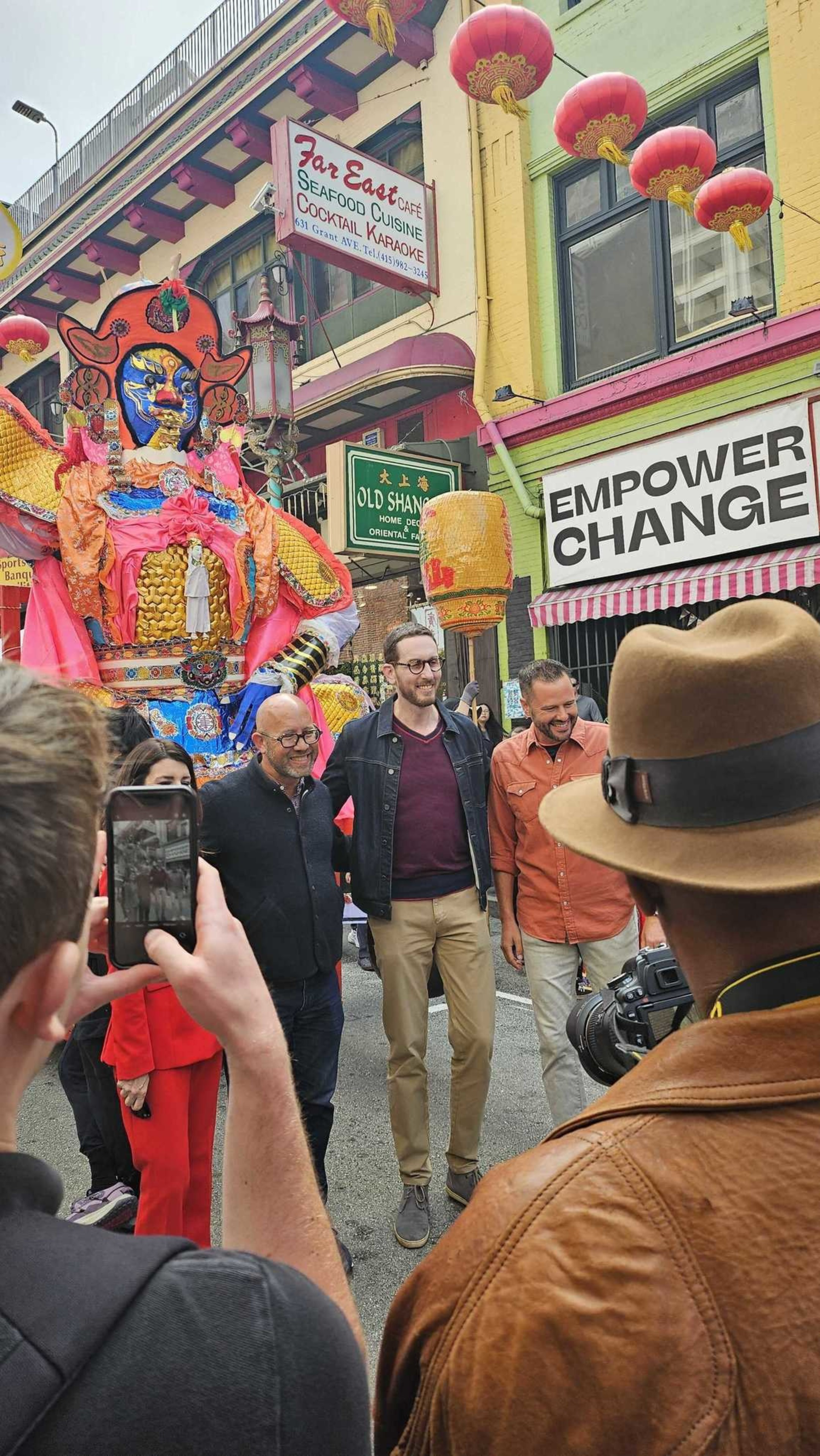 A colorful dragon figure towers behind three smiling men posing on a bustling street decorated with red lanterns and vibrant signs.