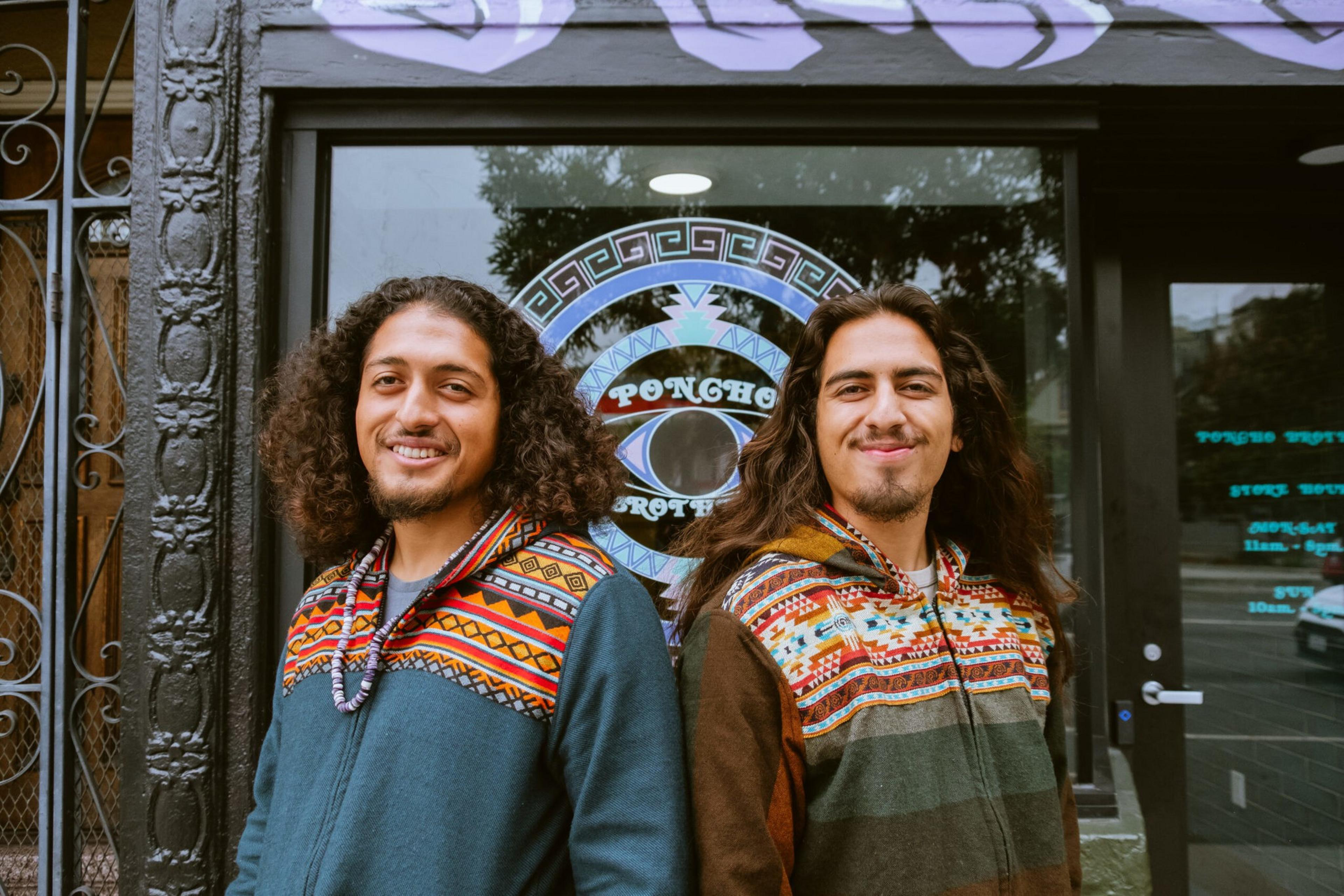 Two men with long hair smile, standing back-to-back in colorful patterned hoodies in front of a glass door with a circular logo.