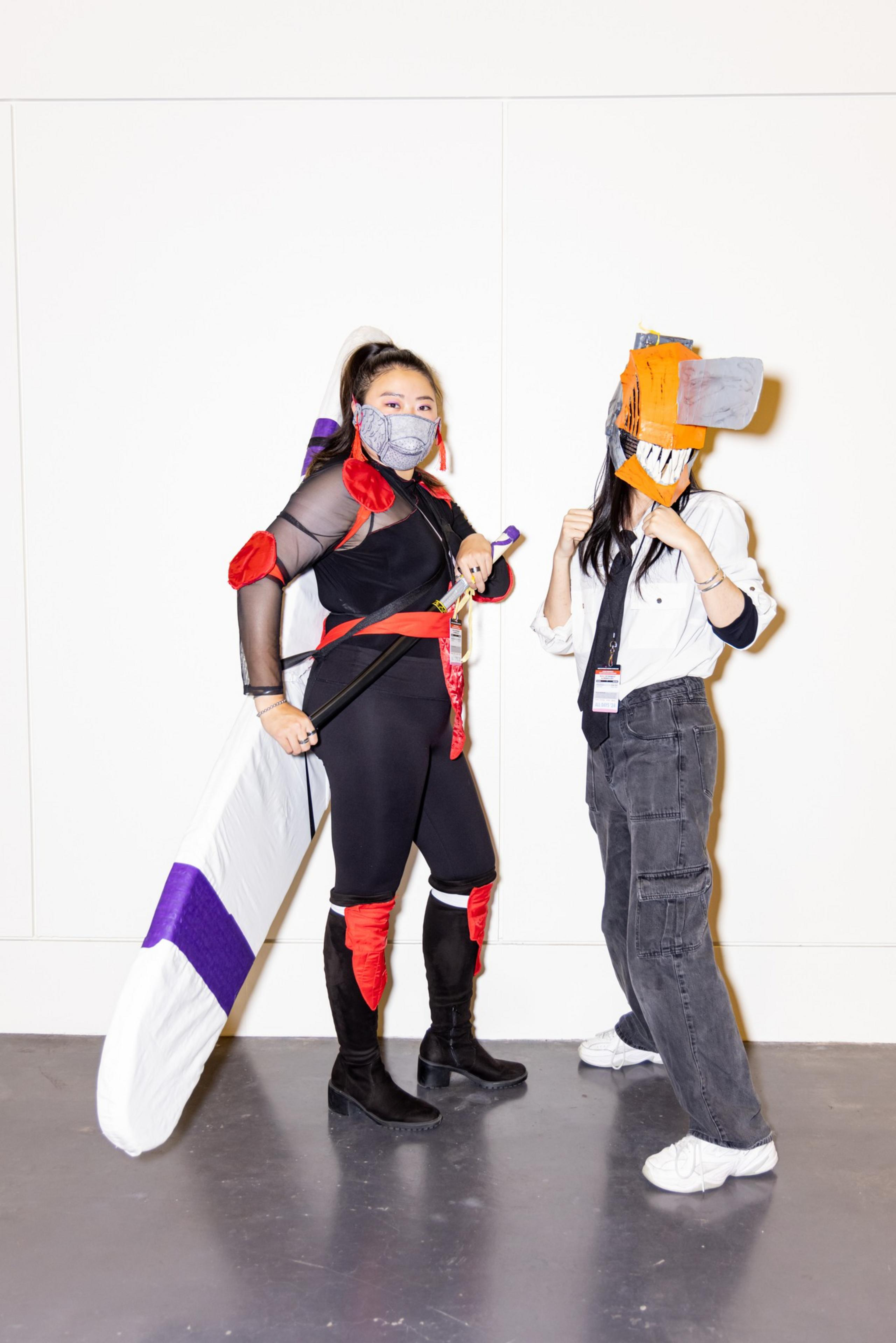 Two people in cosplay pose for a photo. One wears a ninja-like outfit with a mask and large sword, while the other wears a shirt, tie, and headgear resembling a chainsaw.