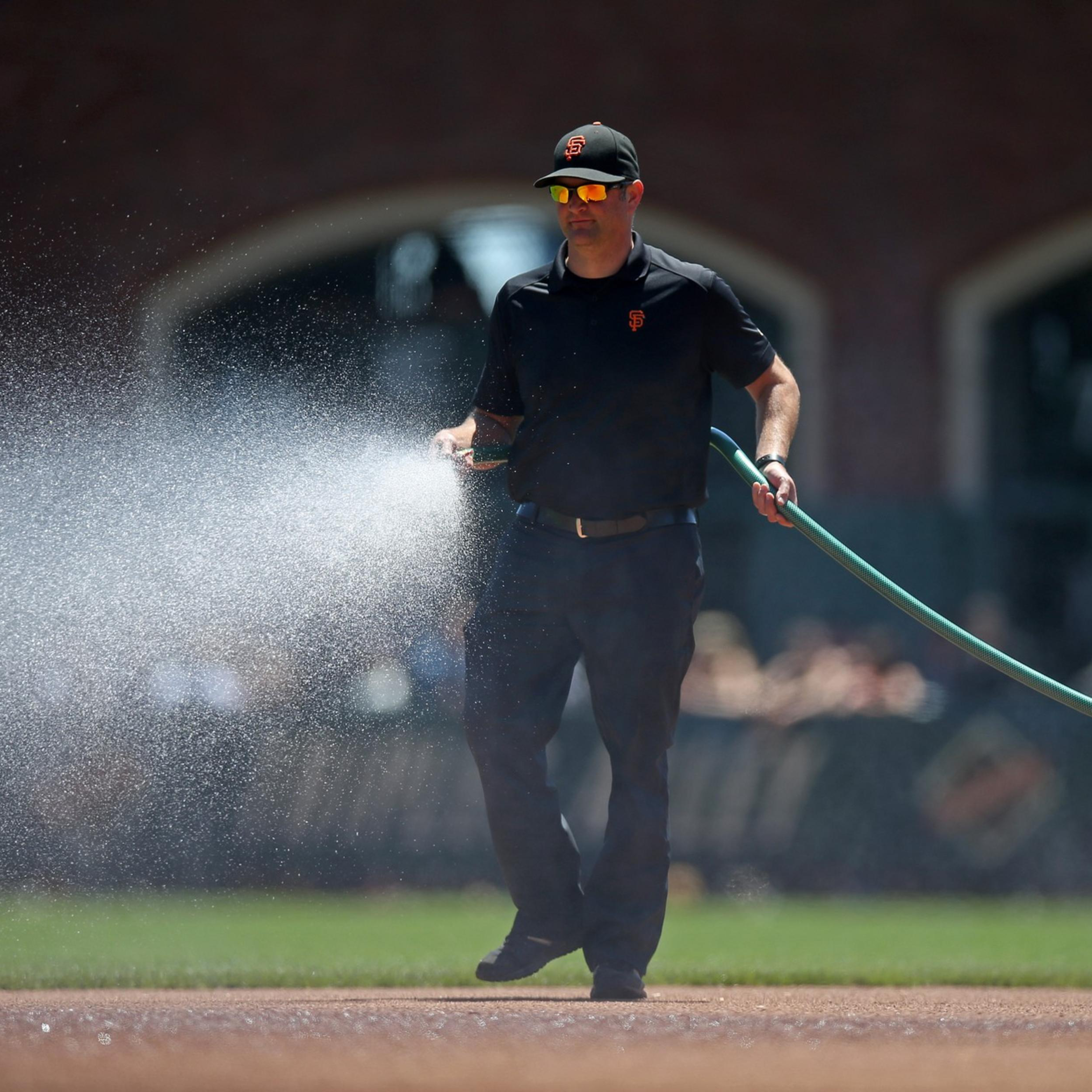 A man wearing a black outfit and cap waters a baseball field with a green hose, spraying water over the dirt area.