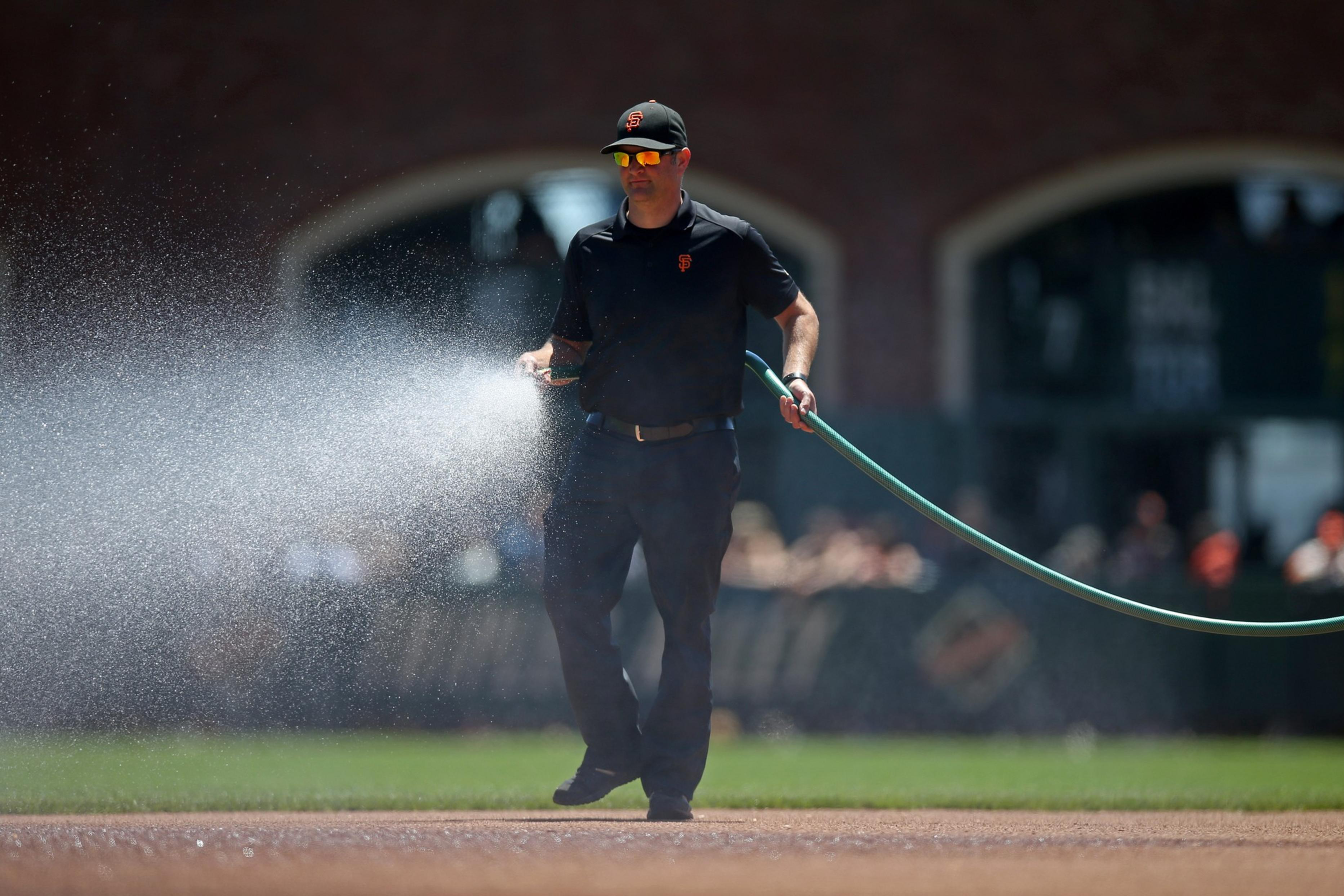 A man wearing a black outfit and cap waters a baseball field with a green hose, spraying water over the dirt area.