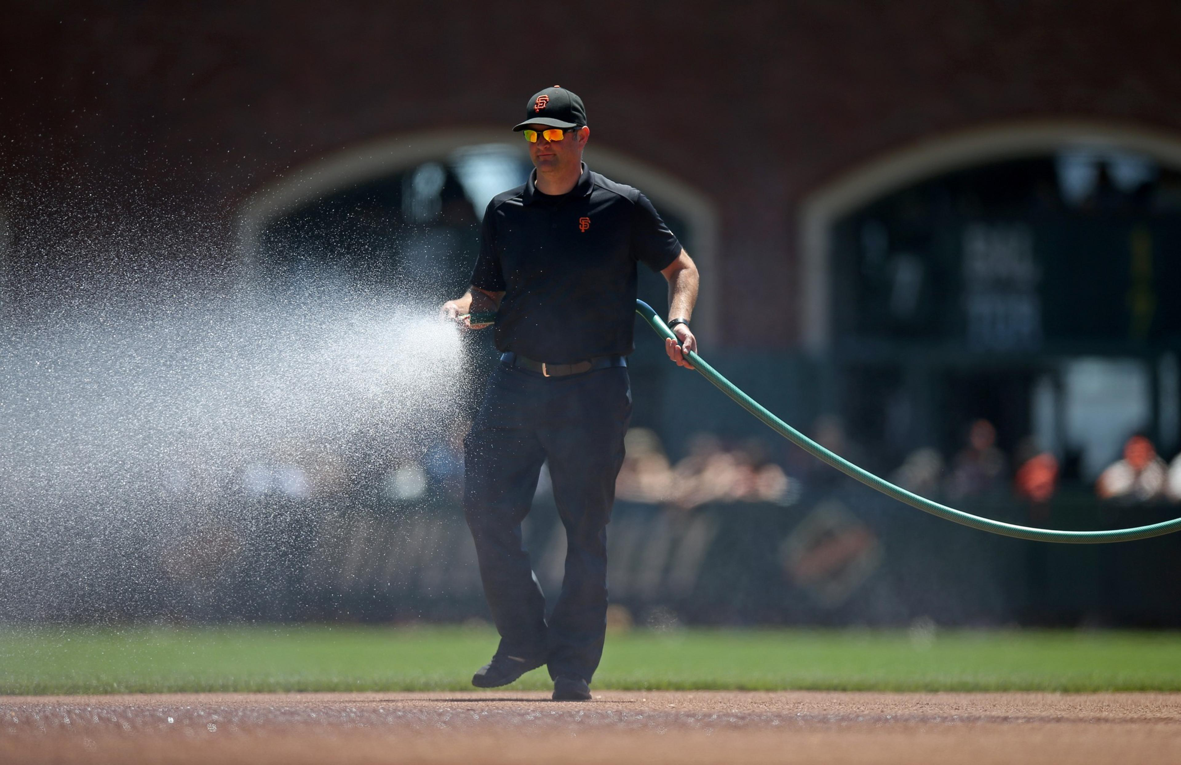 A man wearing a black outfit and cap waters a baseball field with a green hose, spraying water over the dirt area.