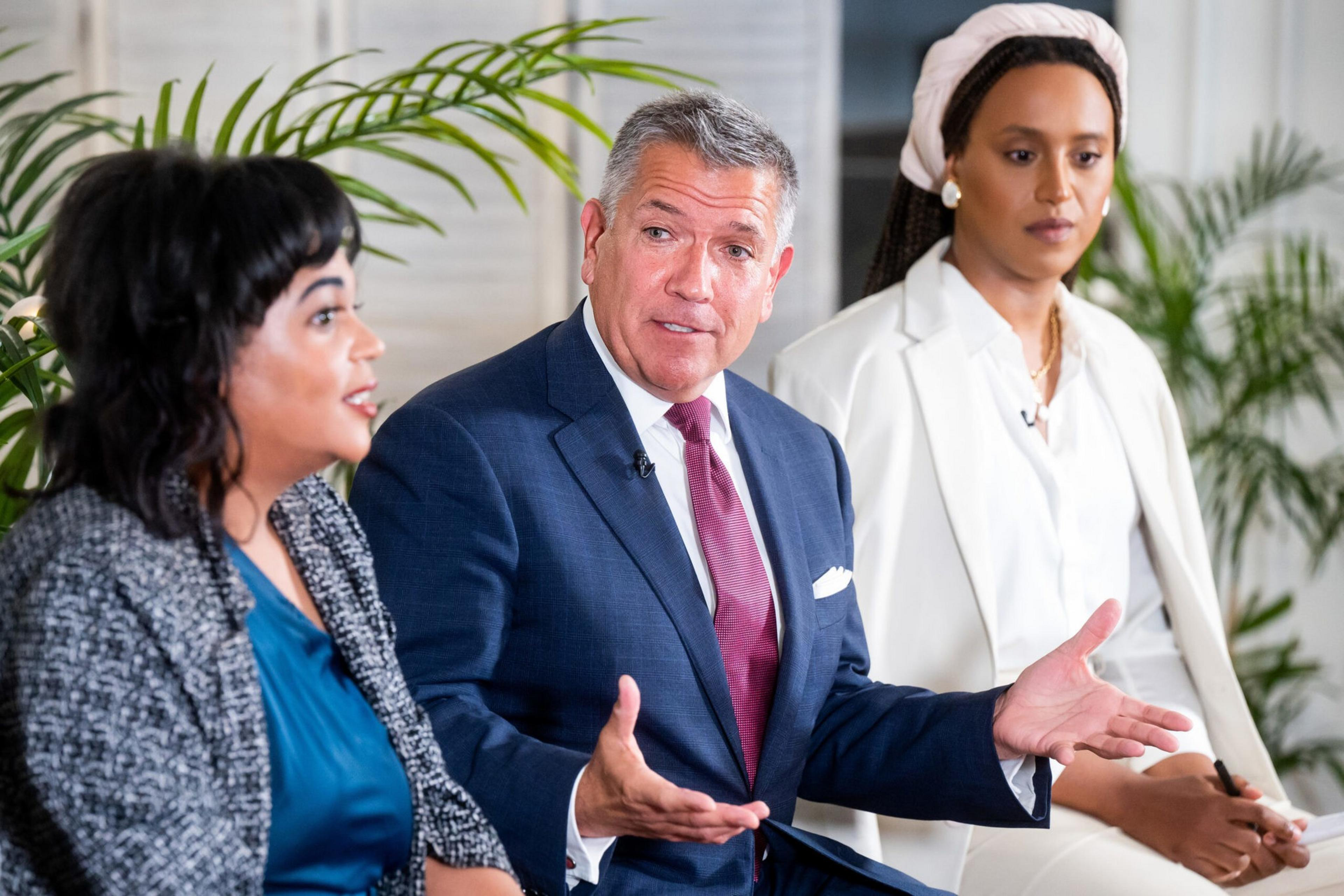 A man in a blue suit and red tie gestures while speaking between two women, one in a gray jacket and blue top, the other in an all-white outfit with a headband.
