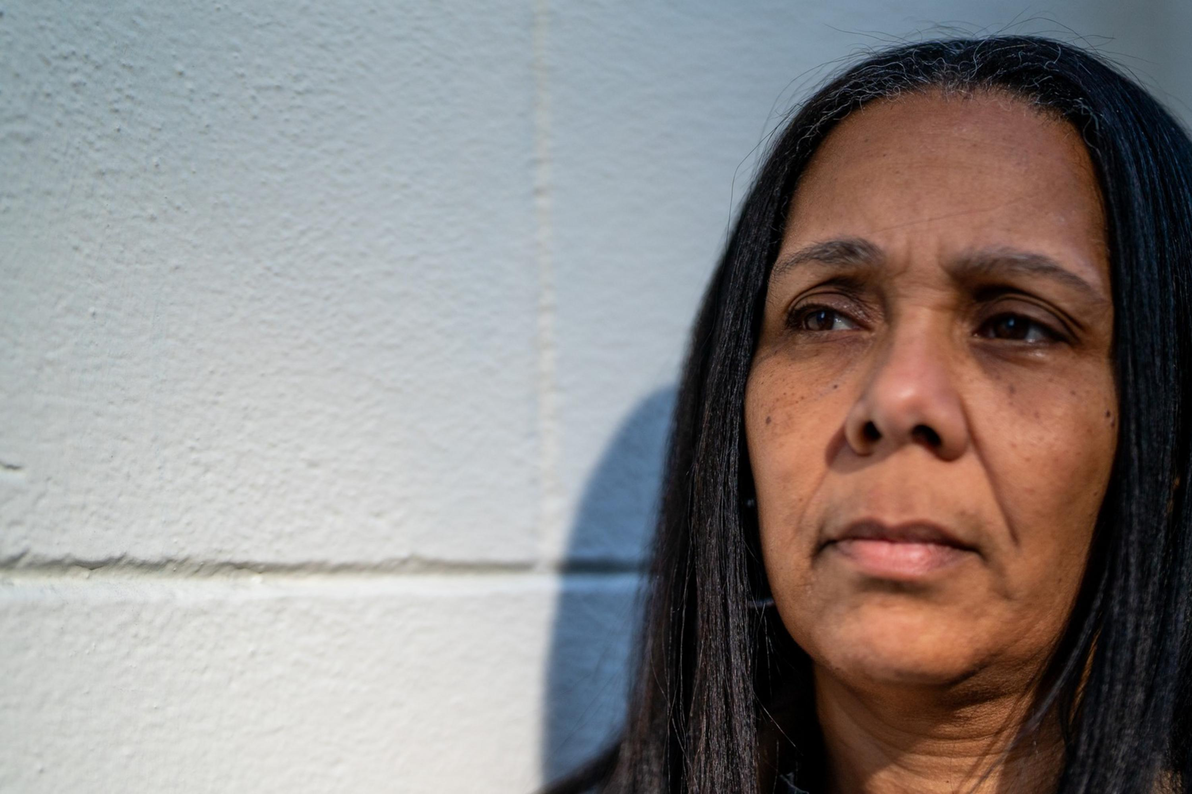 A woman with long dark hair stands against a light-colored wall, looking thoughtfully to the side. She's wearing a black top, and sunlight illuminates her face.