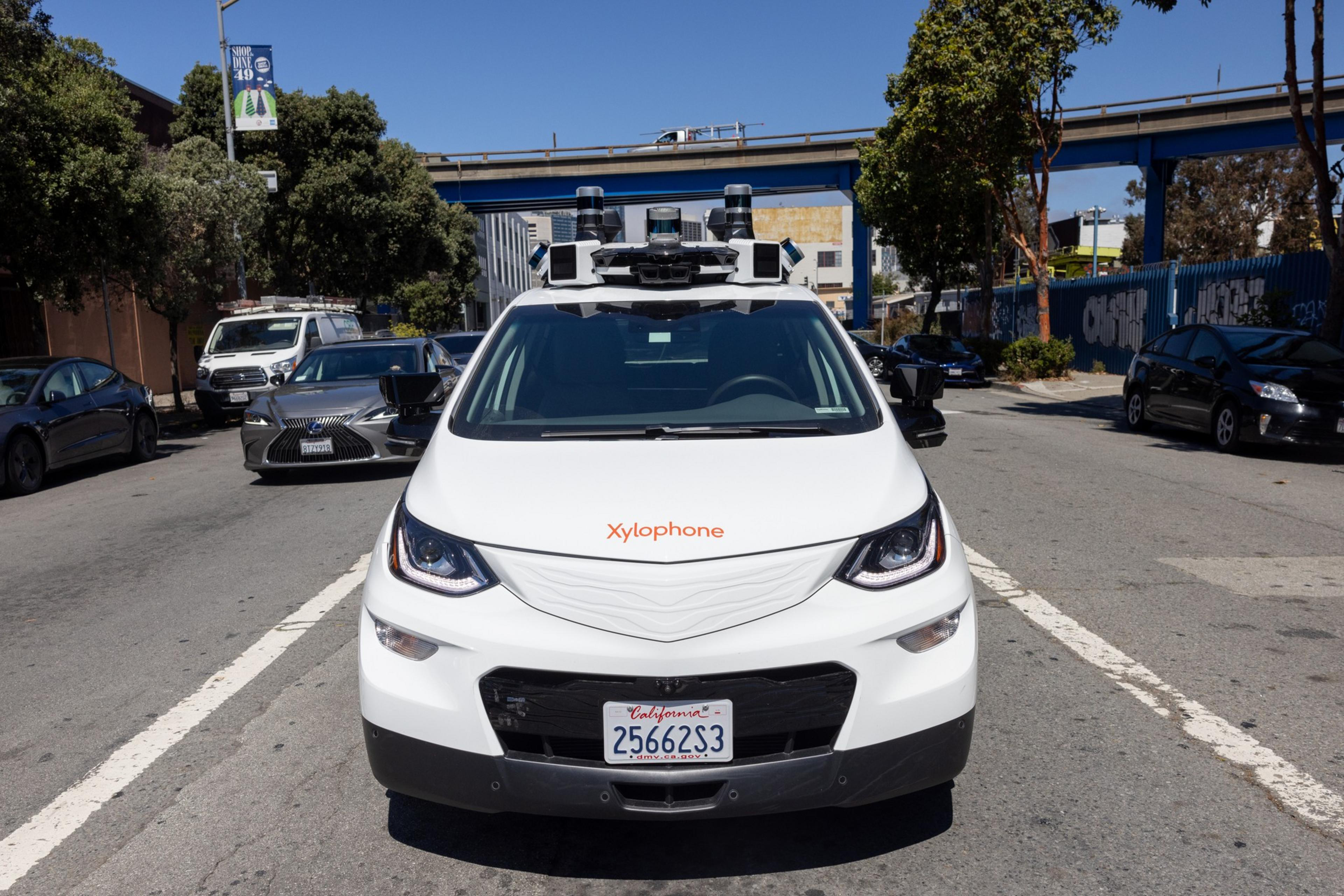 A white car with "Xylophone" on the front, equipped with sensors on the roof, is parked on a city street with other cars and trees around.