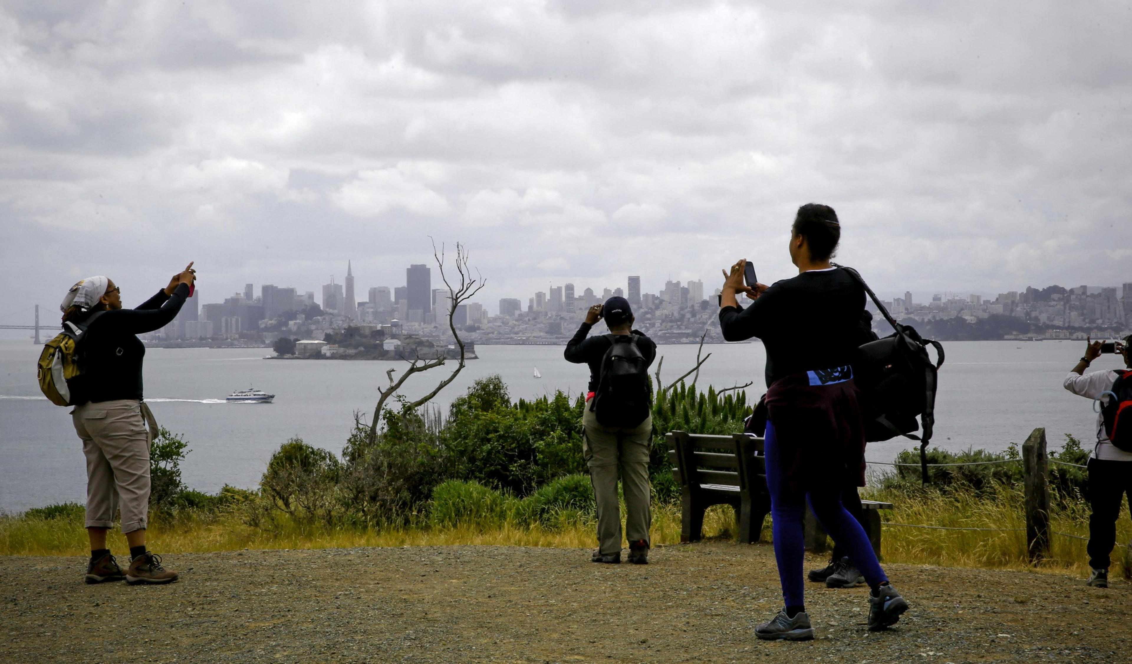 Several people are taking photos with smartphones against a city skyline, with water and a boat visible in the foreground.