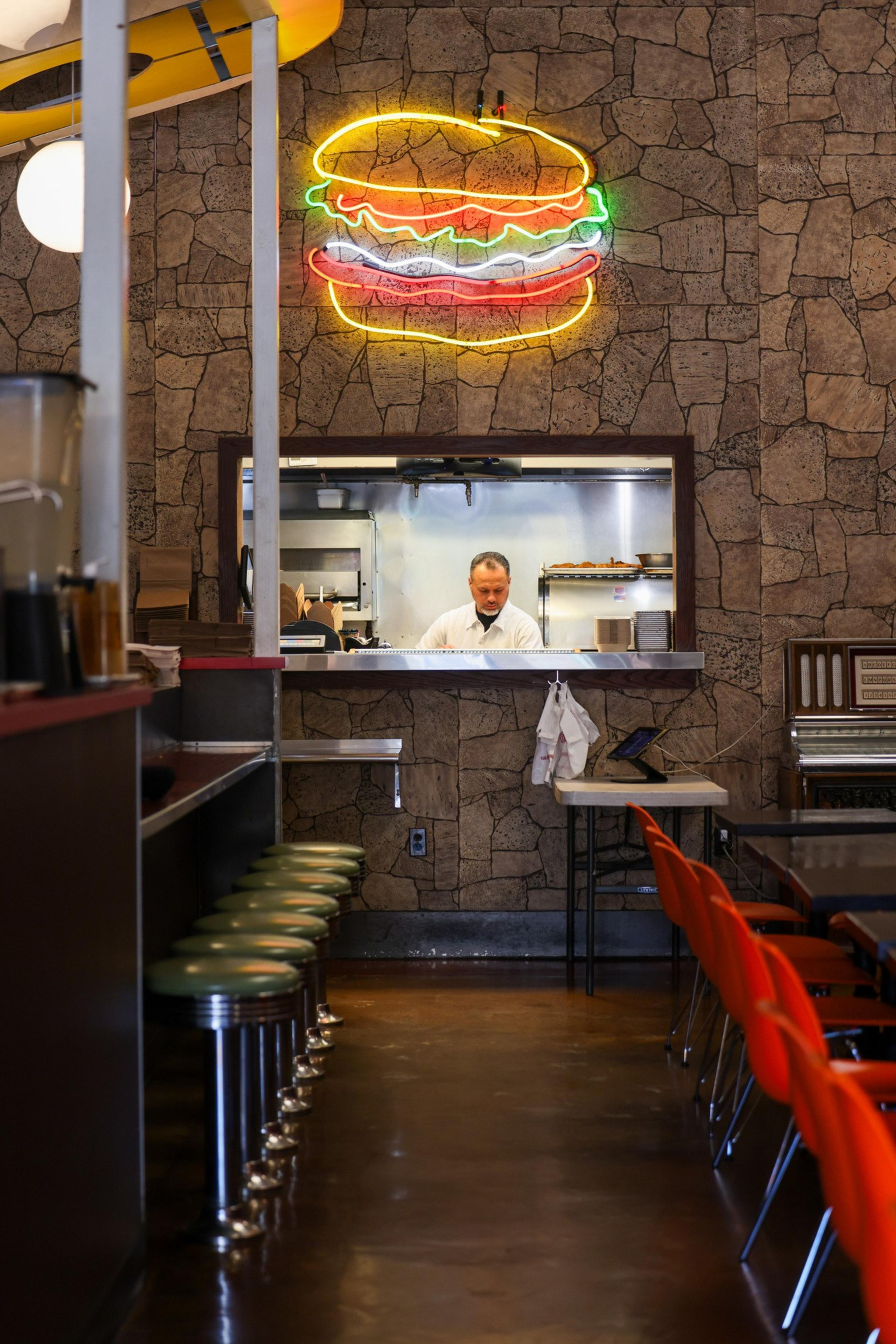 A diner with a stone wall features a neon burger sign. A counter with green stools lines one side, and a man is seen working in the kitchen window.