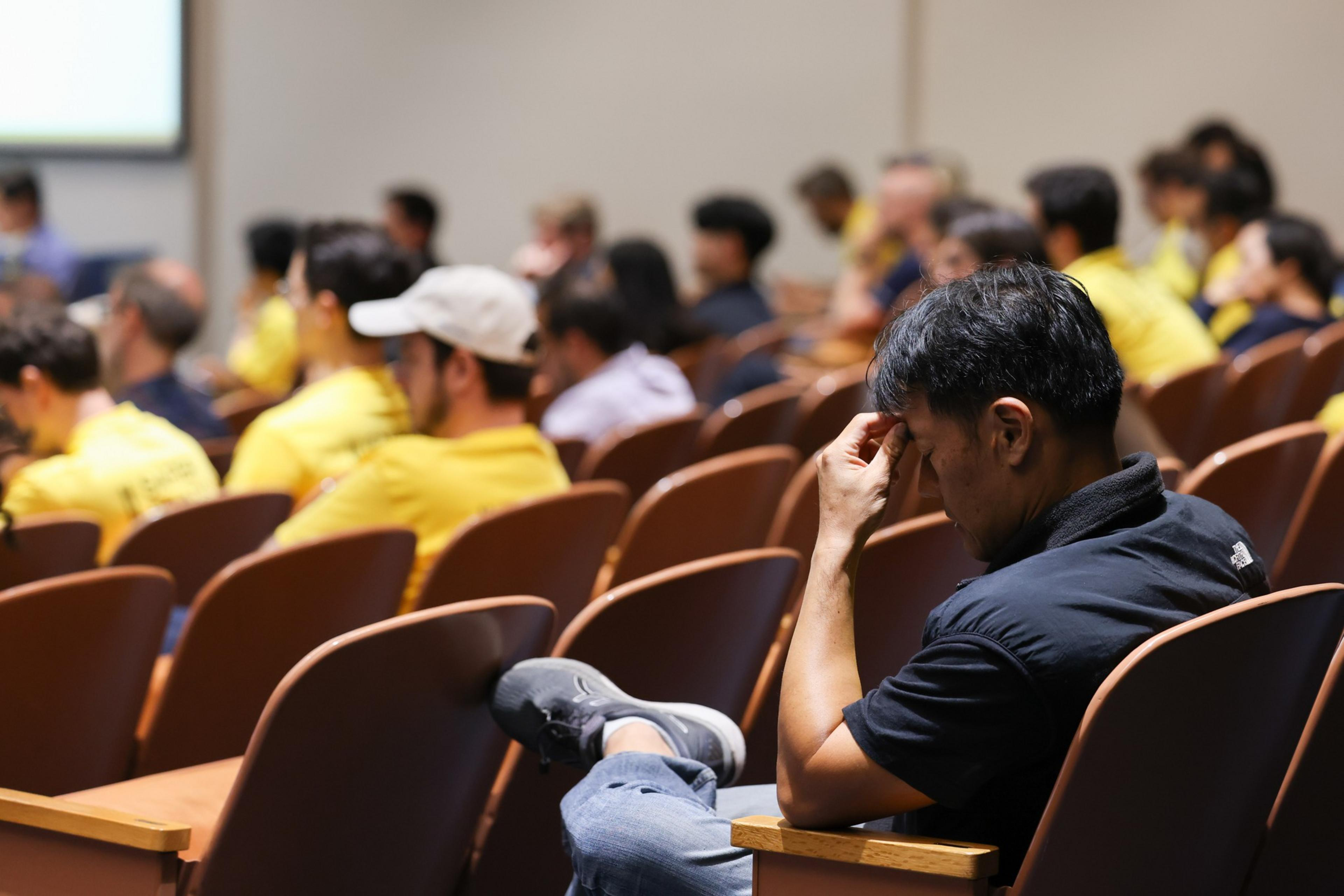A man in a black shirt sits alone with his leg crossed, resting his forehead on his hand, while an audience in yellow shirts listens ahead.