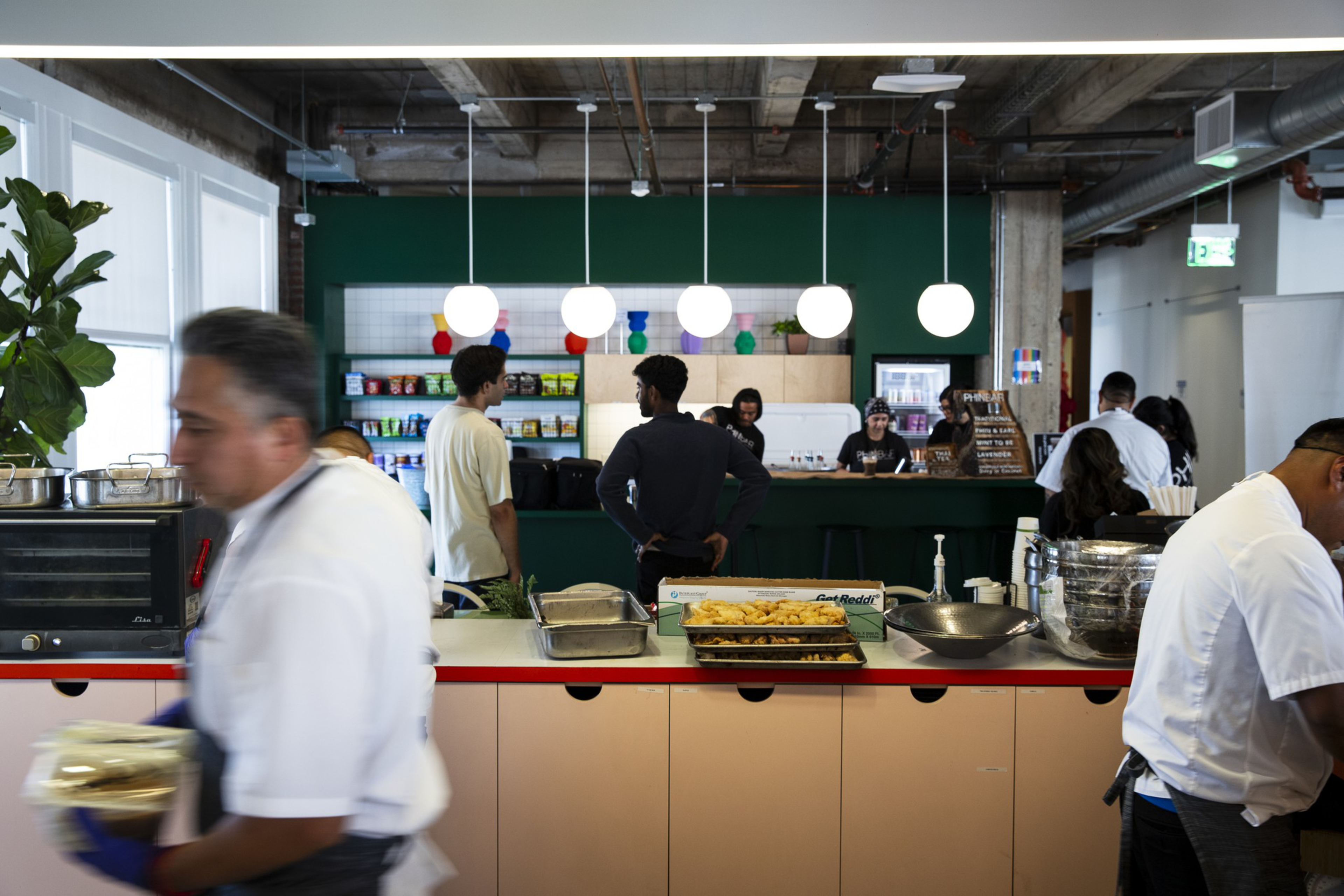 A cafeteria with staff in white uniforms serving food and customers standing at the counter, with hanging globe lights and shelves of snacks in the background.