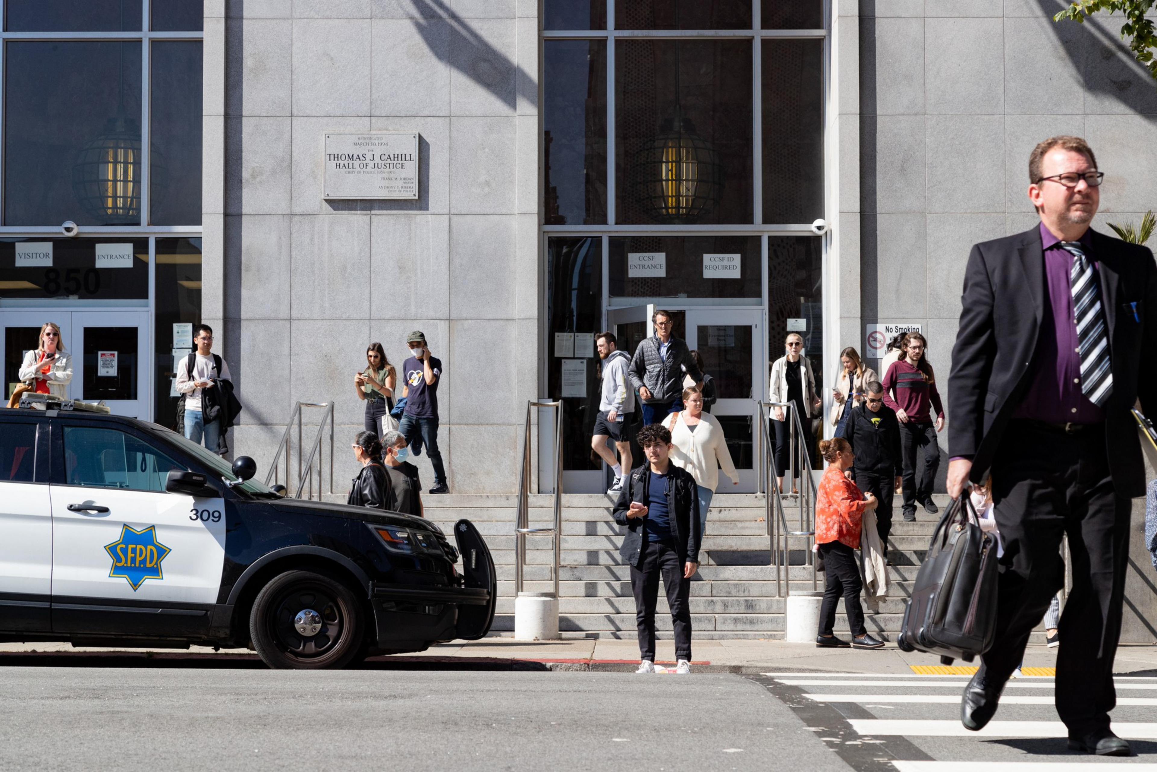 People walk on a sunny street outside a large building.