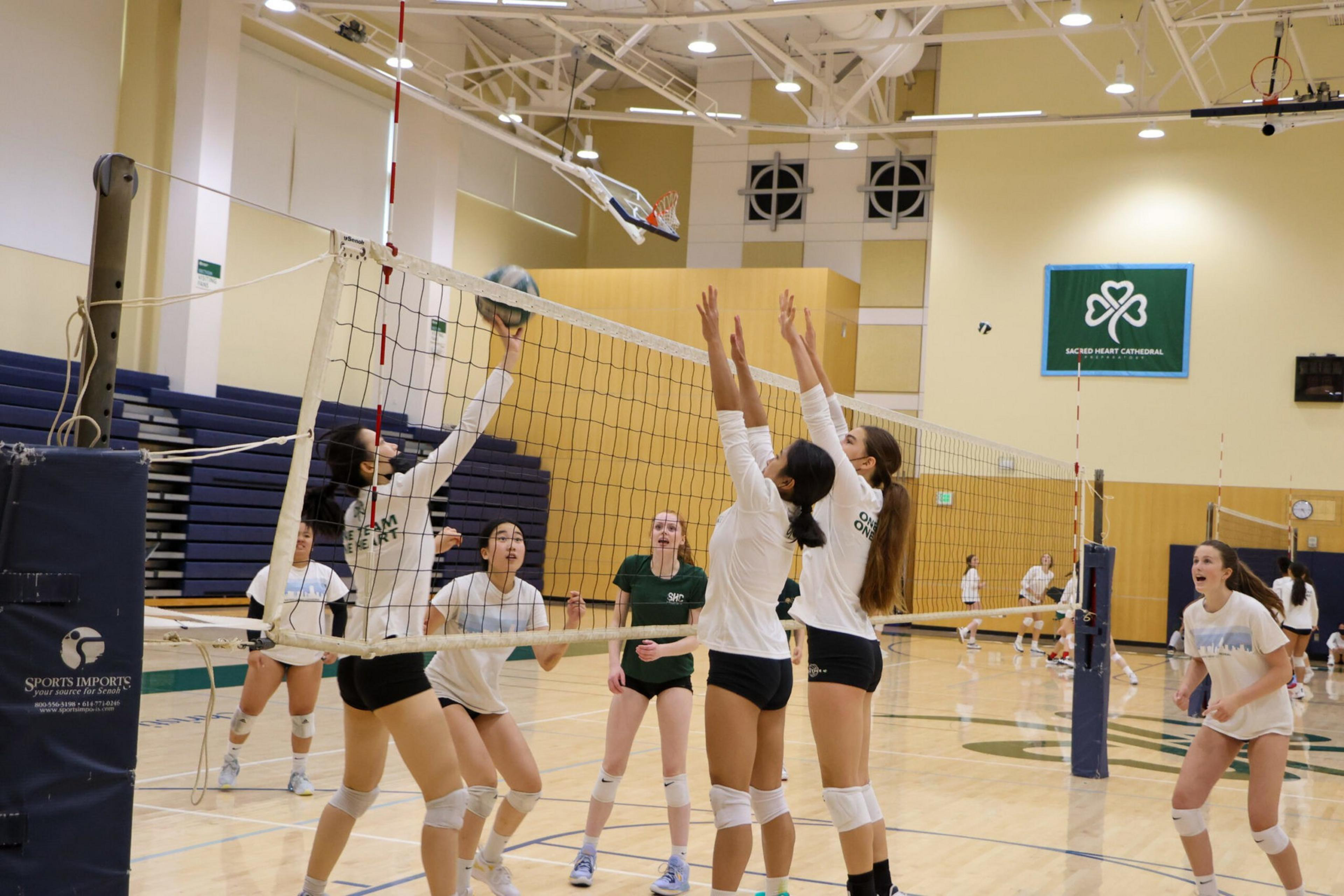 A volleyball game is in progress with players jumping to block or hit the ball over the net inside a gymnasium.