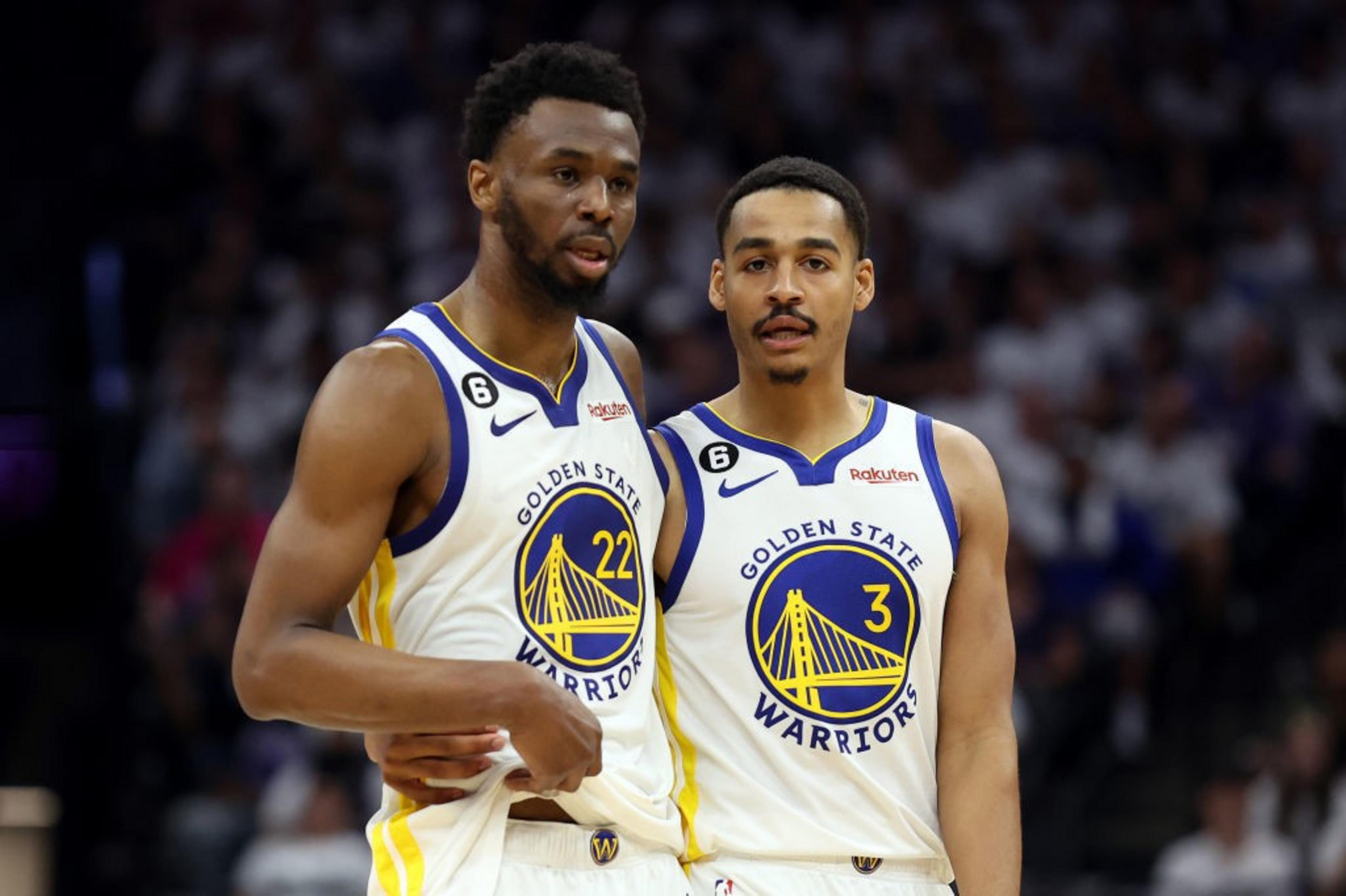 Two basketball players from the Golden State Warriors are on the court. One has his arm around the other's shoulder. Both wear white jerseys with blue and yellow details.