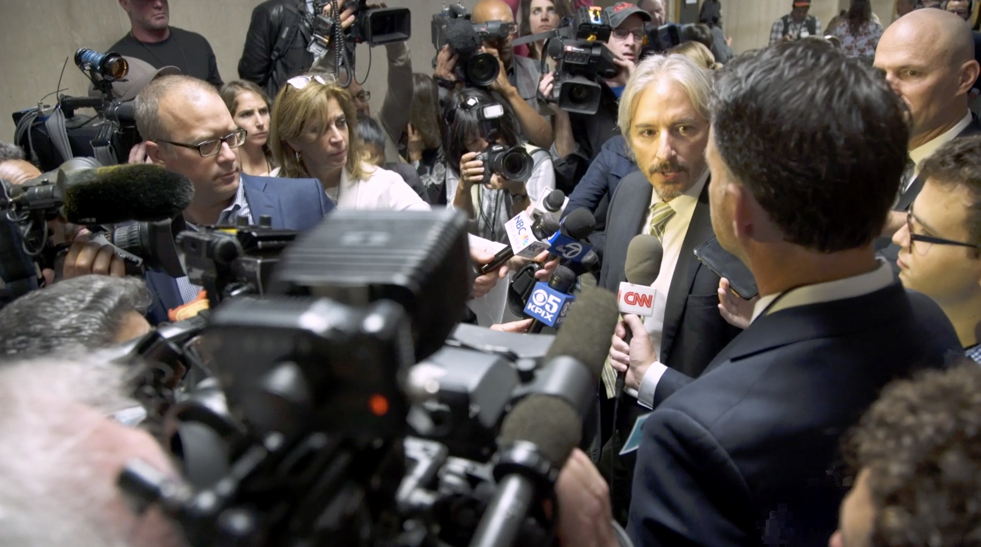 A crowded press conference features numerous reporters with microphones and cameras, surrounding a man in a suit speaking to the media.