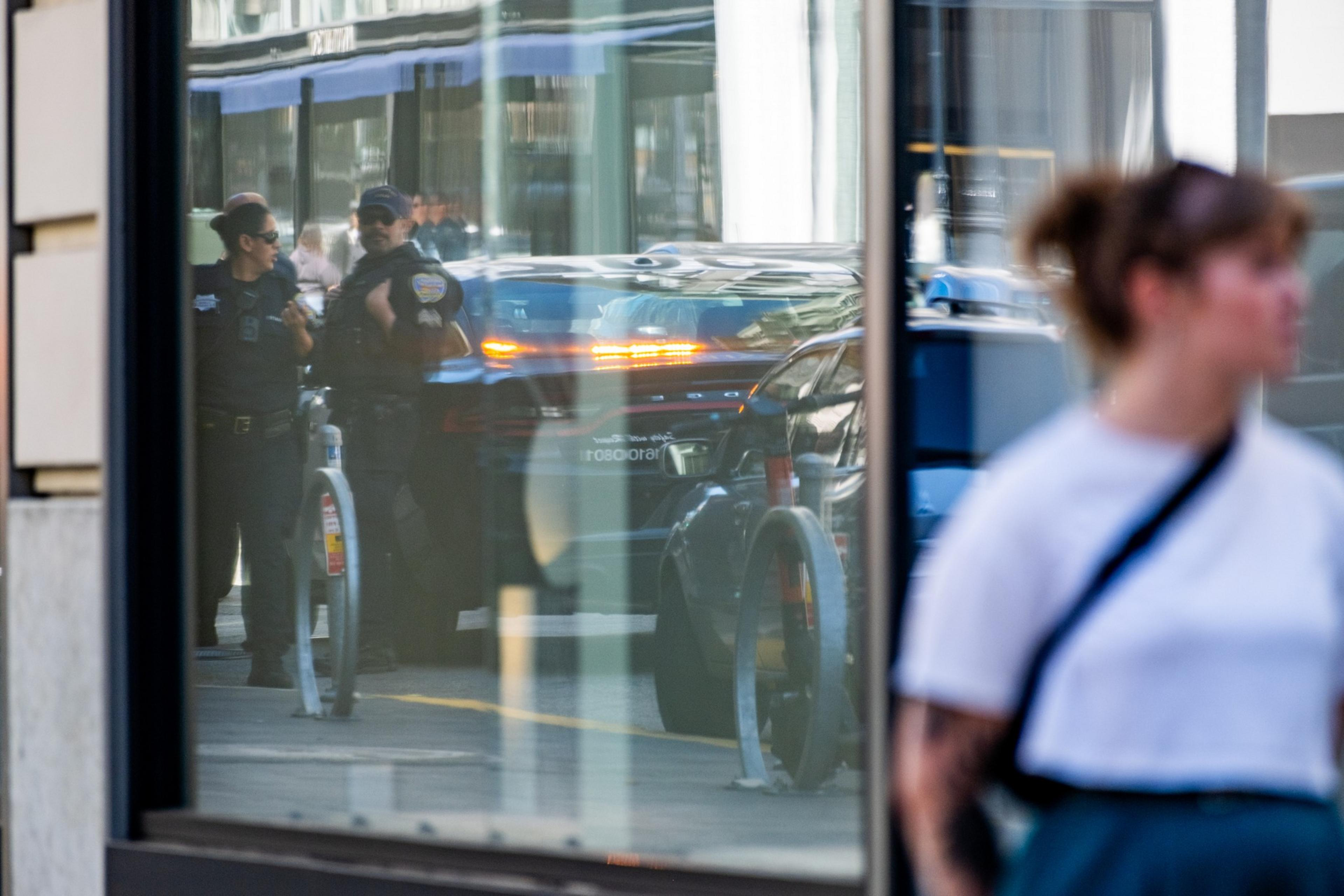 A blurred woman in the foreground appears to pass by a window showing reflections of two police officers, their patrol car, a bus, and part of a bike rack.