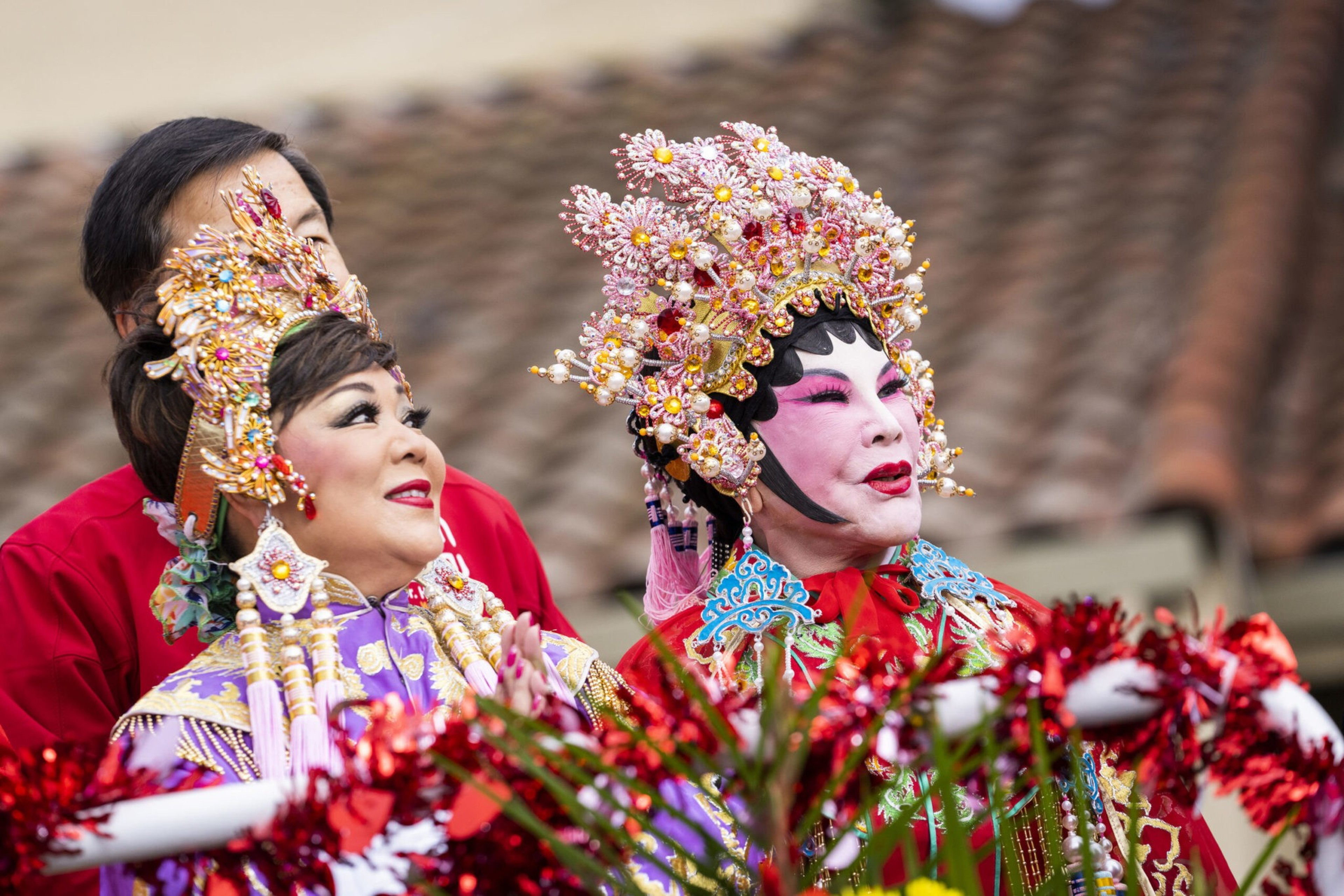 Two performers wear elaborate traditional costumes and floral headpieces, with bright makeup and decorative accessories.