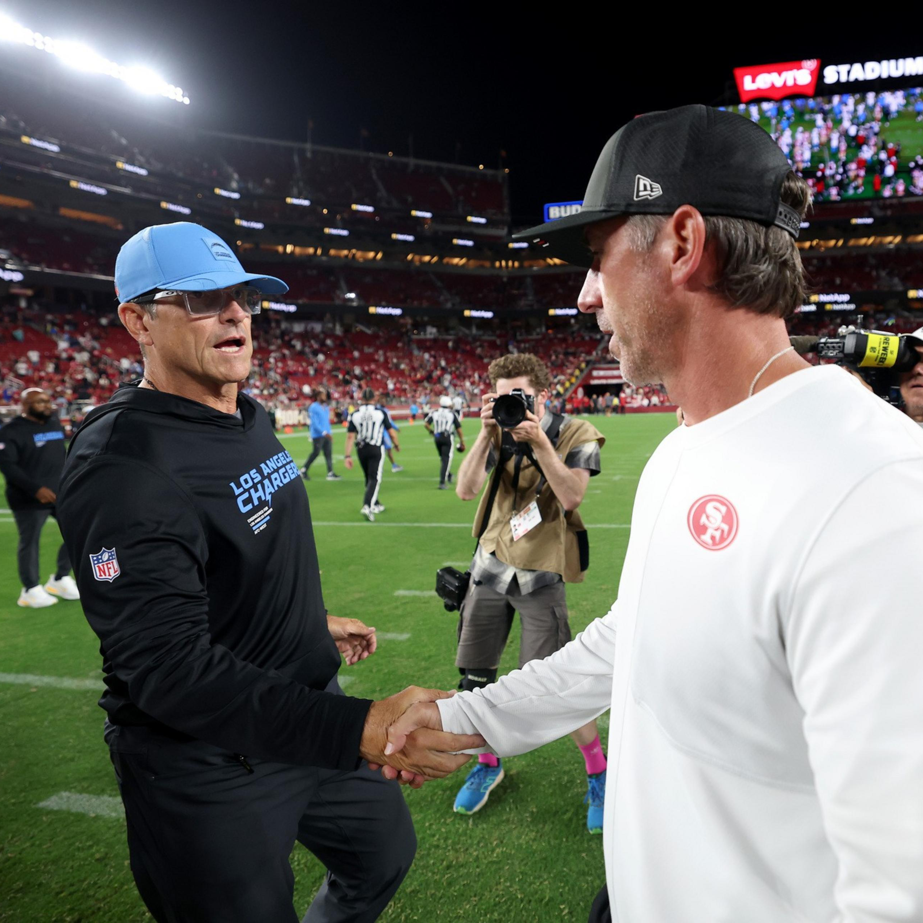 Two NFL coaches, one wearing Los Angeles Chargers gear and the other in San Francisco 49ers gear, shake hands on a football field at Levi's Stadium.