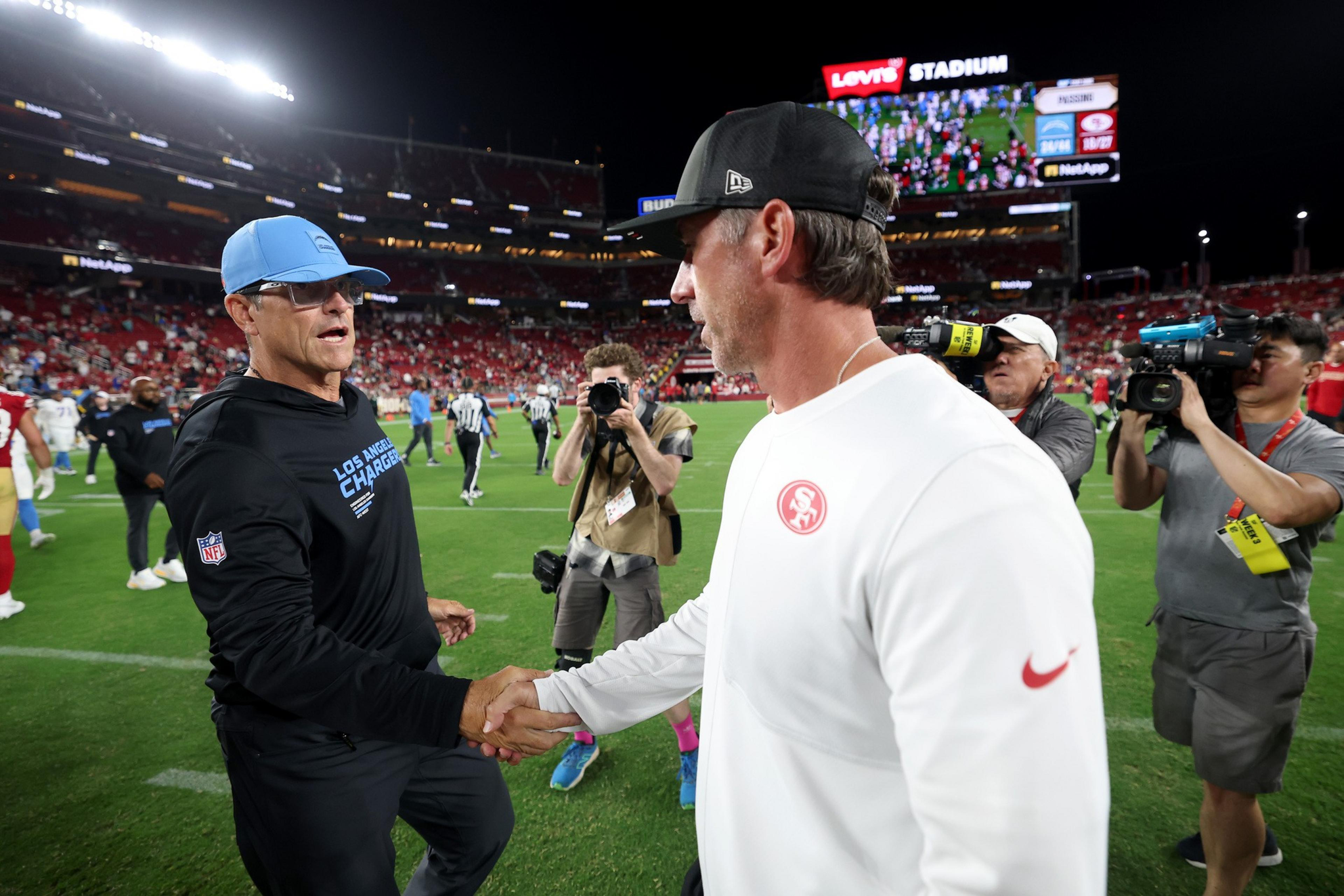 Two NFL coaches, one wearing Los Angeles Chargers gear and the other in San Francisco 49ers gear, shake hands on a football field at Levi's Stadium.