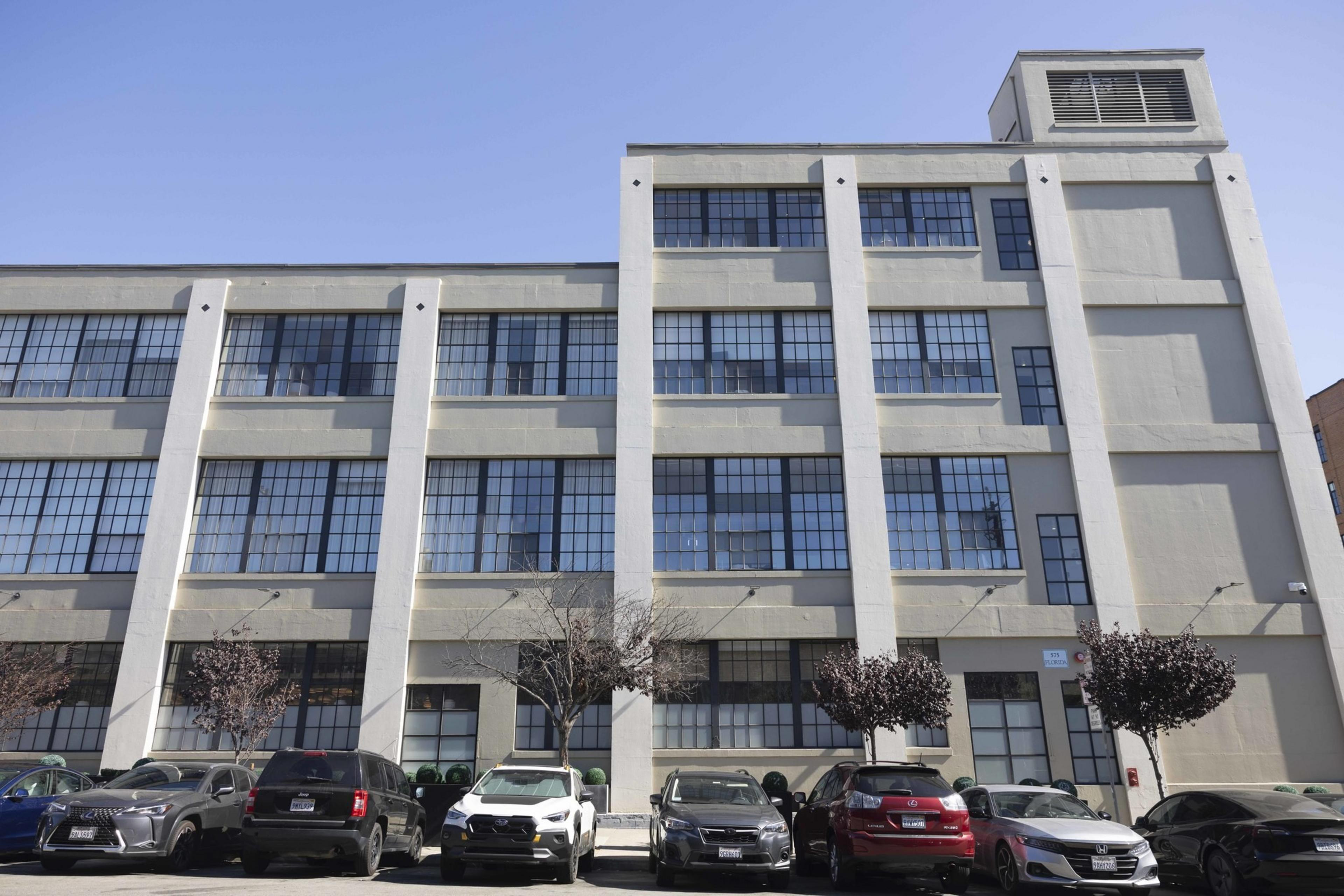 A three-story industrial building with large grid-style windows, set against a clear blue sky, has cars parked in front and a few small trees lining the sidewalk.