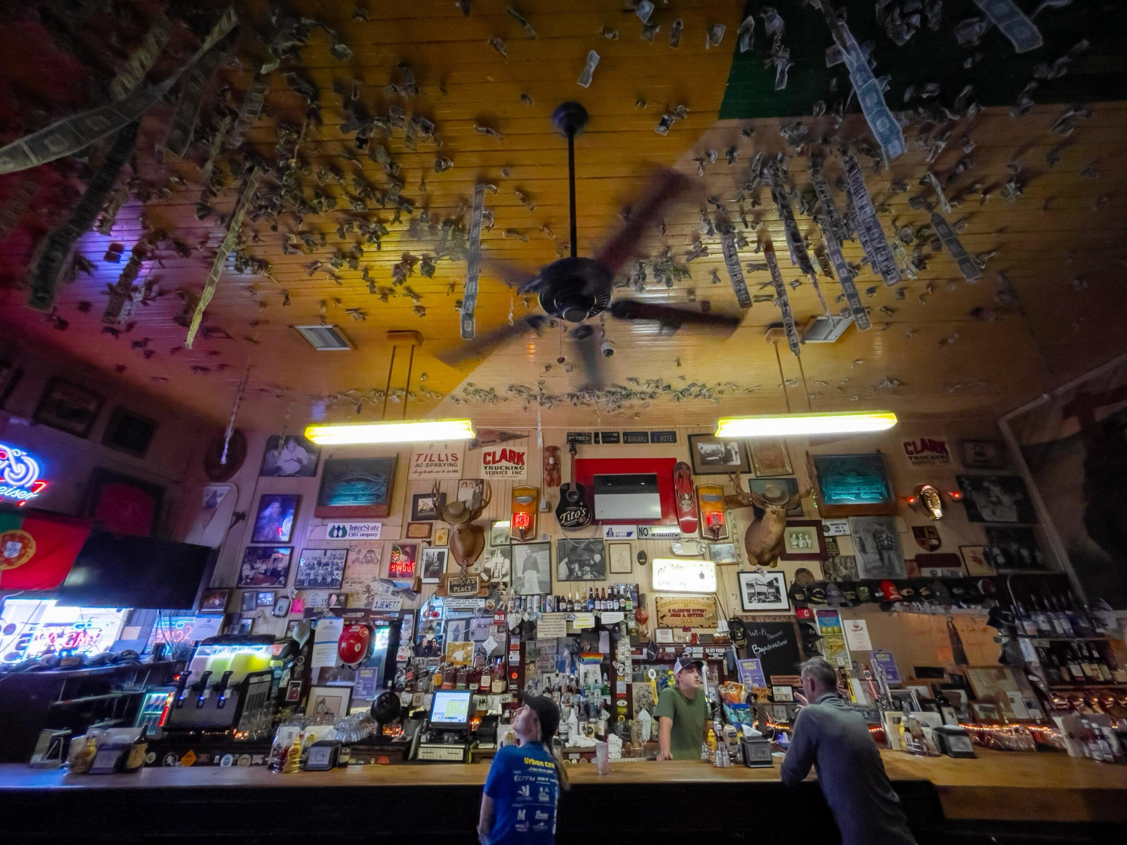 A bar interior with dollar bills attached to the ceiling, three patrons at the counter, mounted deer heads, and a ceiling fan spinning overhead.